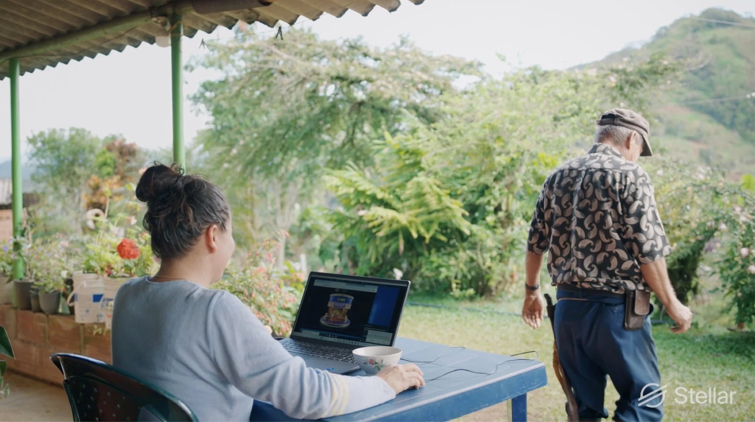 A woman working on a laptop at a blue outdoor table, with a woman in the background holding a knife and gardening outside, surrounded by greenery and flowers.