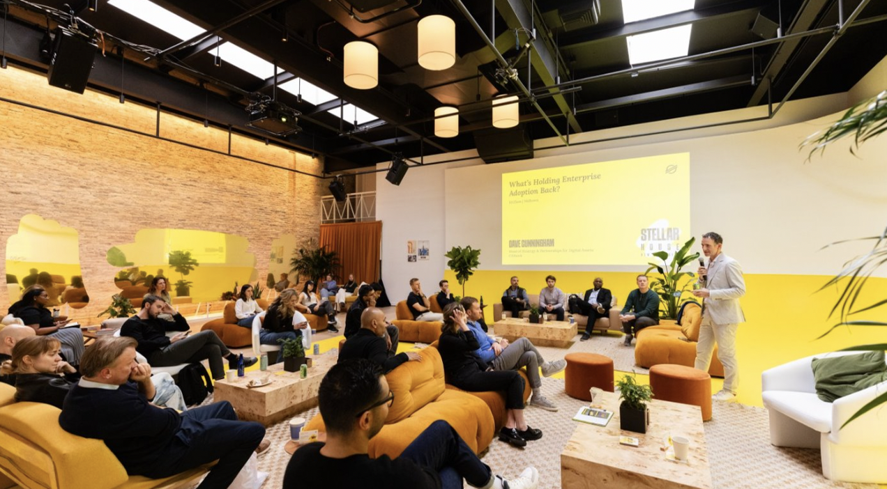A conference or seminar in a spacious room with yellow and white decor. A speaker in a light-colored suit stands with a microphone in front of a large yellow presentation screen, while the audience listens attentively. The room has modern furniture, plants, and a brick wall on one side.