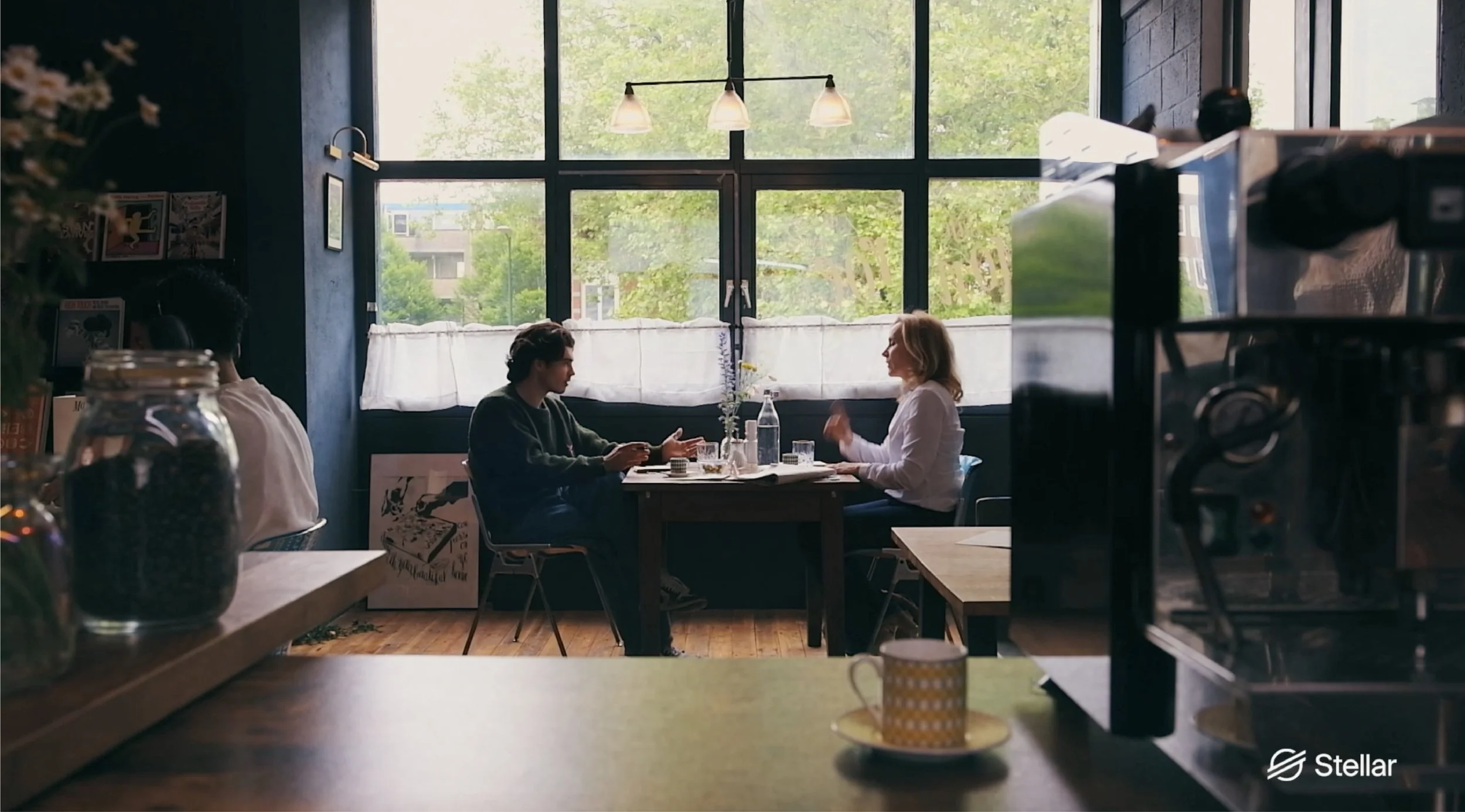 Two people sit across from each other at a table in a bright coffee shop, engaged in conversation, with large windows letting in natural light.