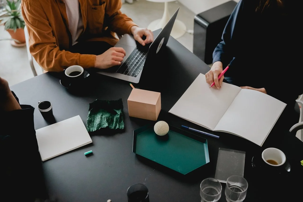 Top-down view of a workspace with two people. One person is typing on a laptop, while the other is writing in an open notebook with a pink pen. The table has various items including cups of coffee, a green tray with a white ball, a pink cube, a piece of black fabric, notebooks, and some glasses of water.