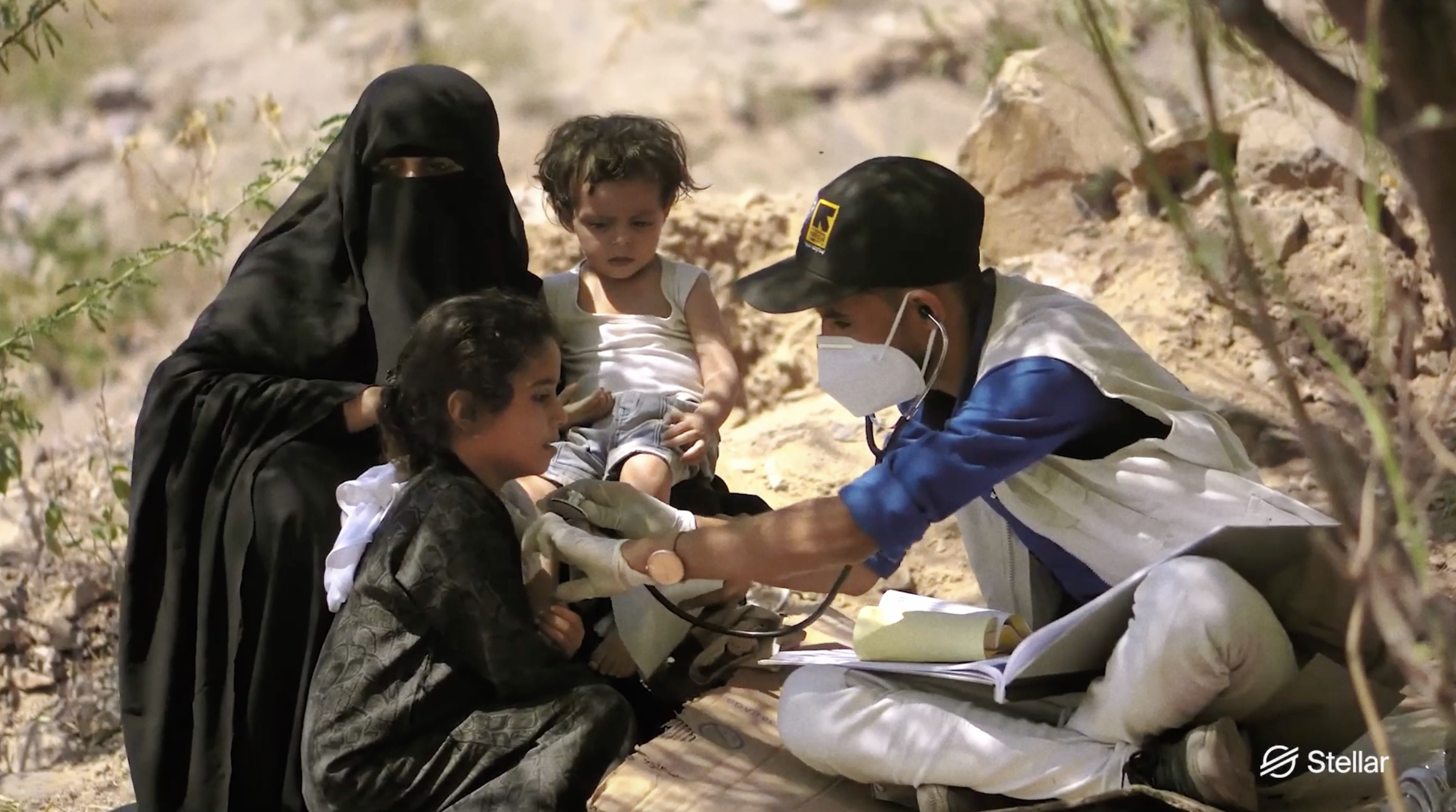 A doctor examines a young girl with a stethoscope while a woman, two children, and a man wearing a face mask and gloves watch outdoors on a rocky terrain.