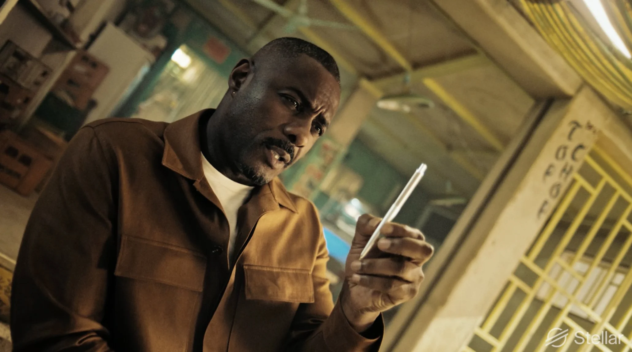 A man in a brown jacket looks at his phone inside a rustic building with yellow and white striped decorations and a partially visible alphabet chart on the wall.