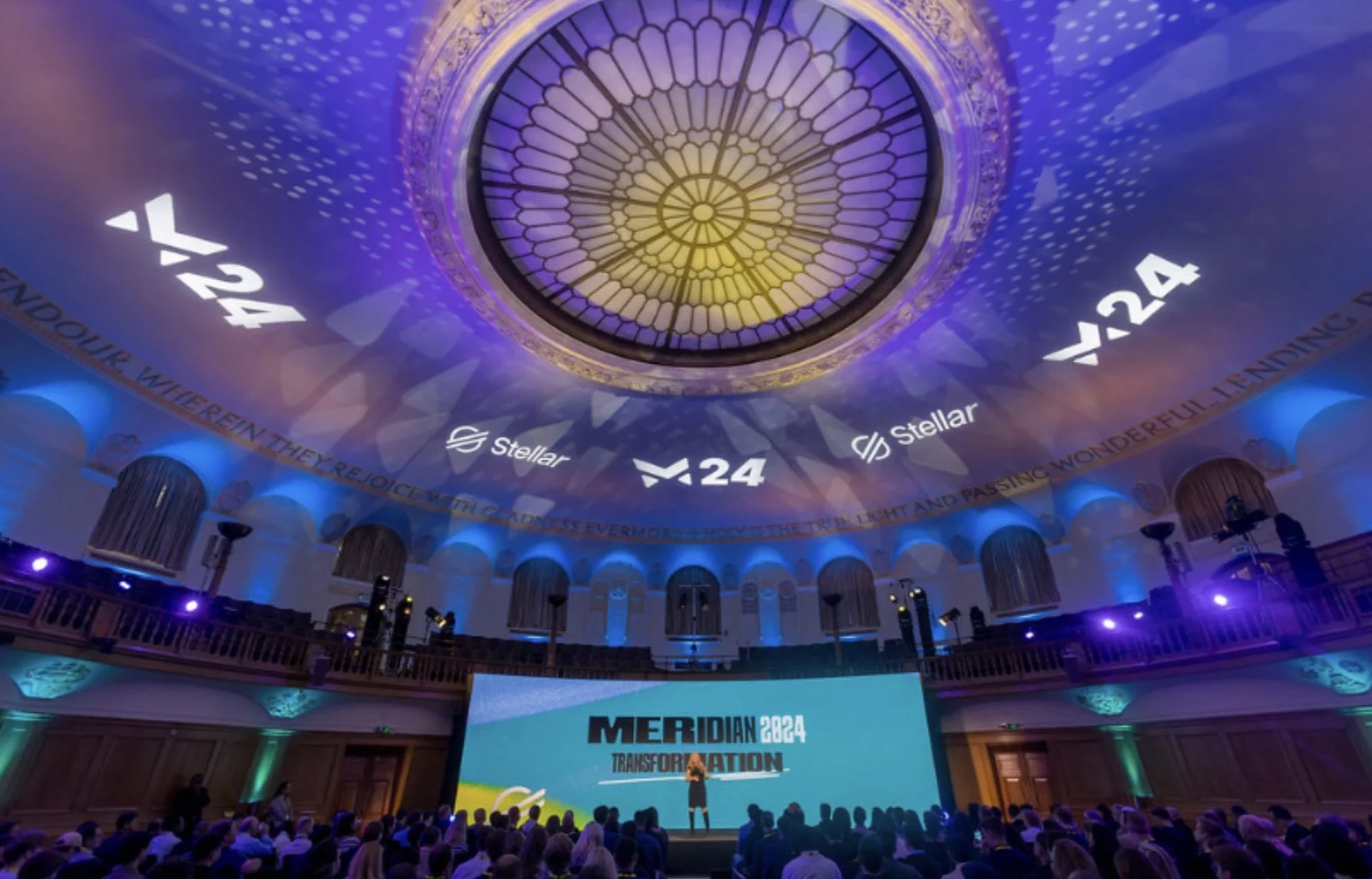 Large conference hall with audience, stage, and a large screen displaying 'MERIDIAN 2024 TRANSFORMATION'. The ceiling has a stained-glass dome with purple and yellow lighting, projections of logos, and numbers 24 and 24:4. The walls are decorated with purple and green lighting.
