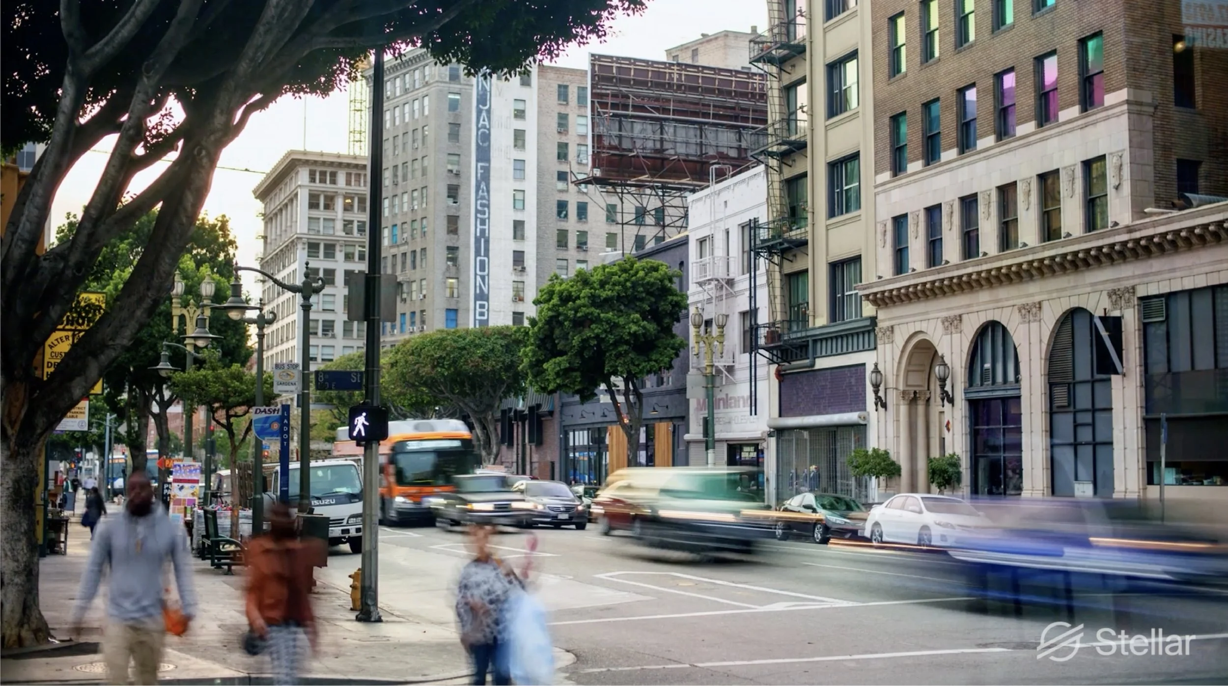 A busy city street scene during daytime with vehicle traffic, pedestrians walking on the sidewalk, and tall buildings in the background.