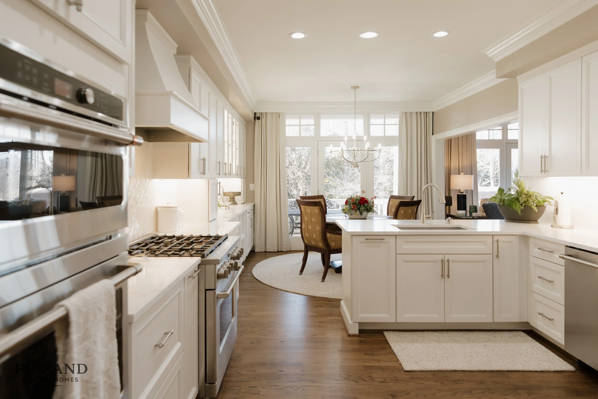 A bright, modern kitchen with white cabinets, a kitchen island, a dining area with table and chairs, large windows with curtains, and a chandelier, overlooking a snowy outdoor scene.