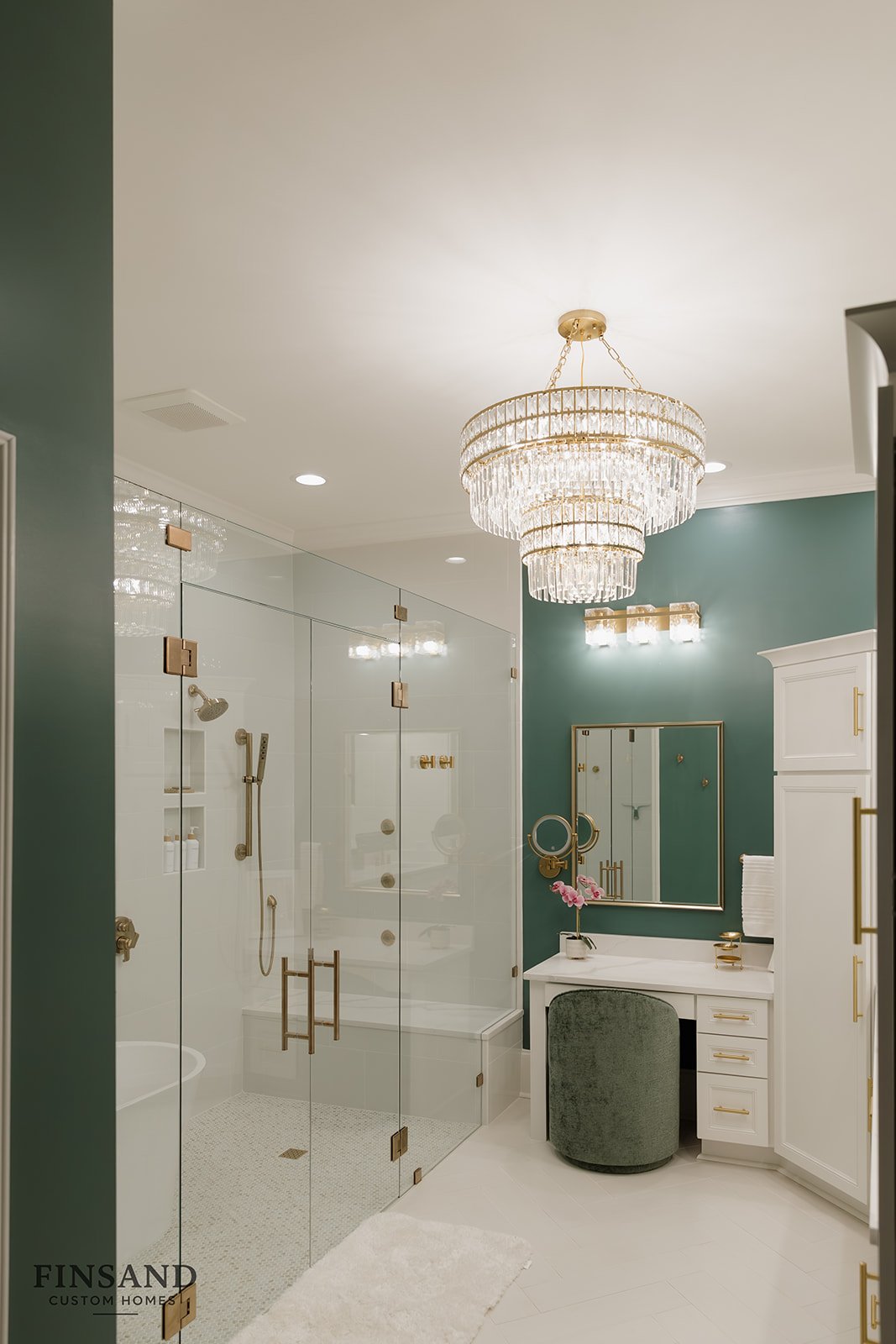 Luxurious bathroom with a glass shower enclosure, gold fixtures, a white vanity with gold accents, a large mirror, a chandelier, and teal walls.
