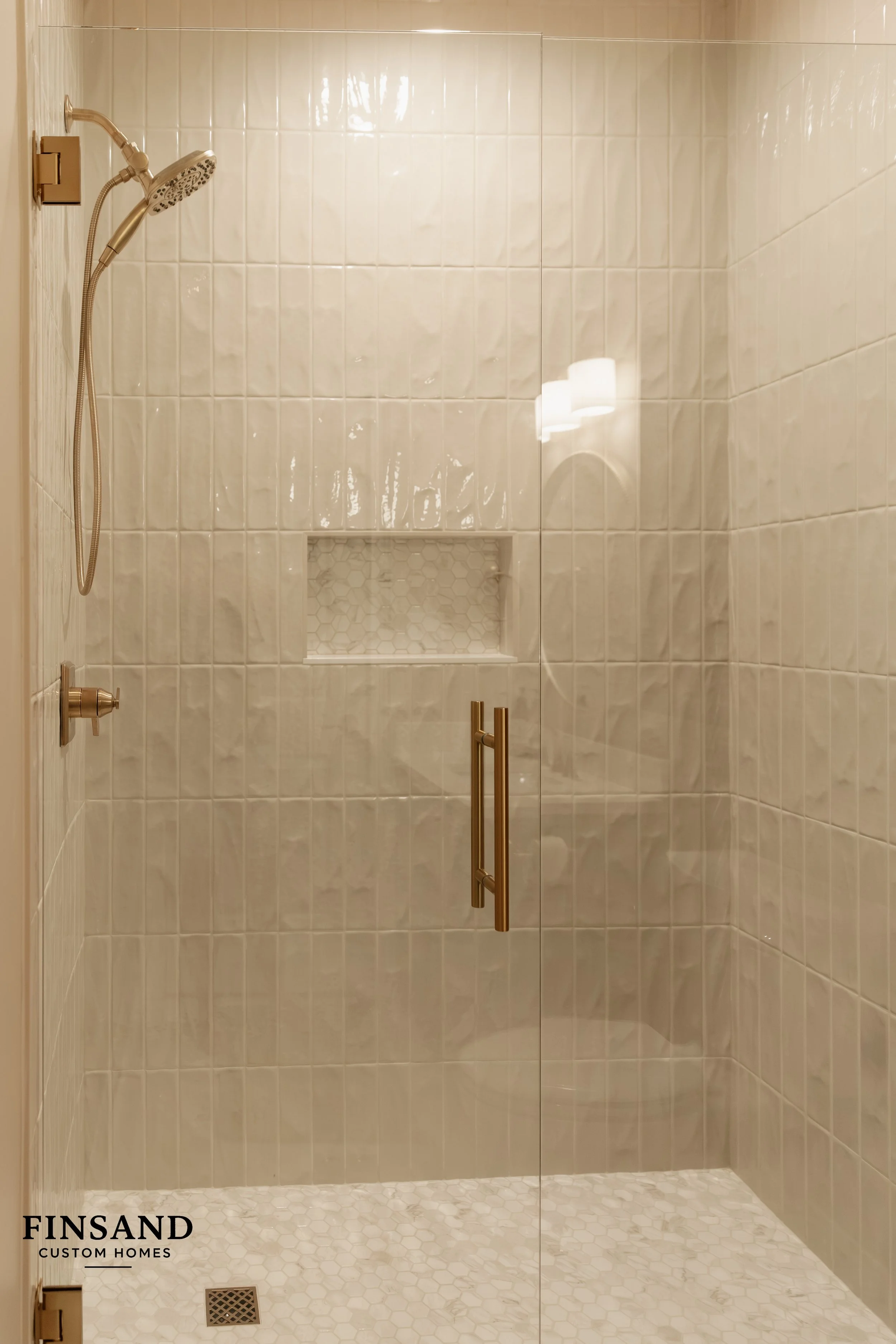 A modern glass-enclosed shower with beige tiles on the walls and small hexagonal tiles on the floor. The showerhead is mounted on the left wall, with a handheld shower attached to a flexible hose. There is a built-in niche with hexagonal tiles for storage. The door has brass handles, and a floor drain is visible at the bottom of the shower."