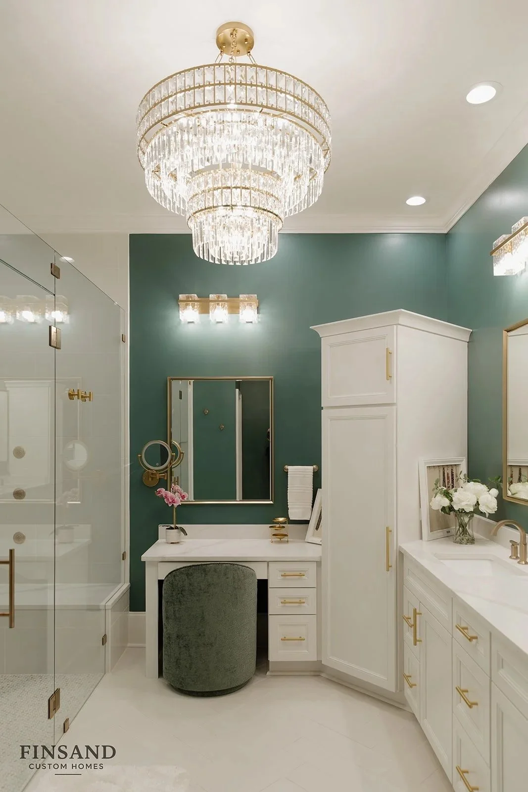 Luxurious Thornblade Greer, SC bathroom renovation with green walls, white cabinetry, gold hardware, a large crystal chandelier, a vanity with a mirror, a white flower arrangement, and a glass shower enclosure.