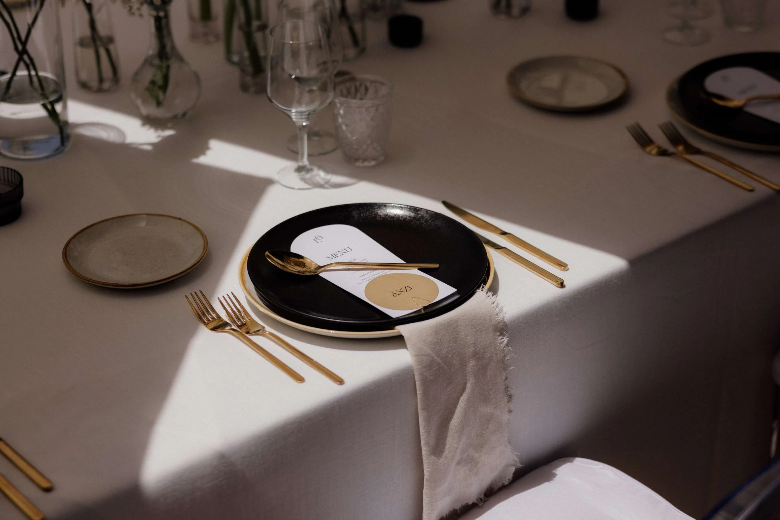 Elegant table setting with a black plate, gold utensils, a white cloth napkin, and a menu card on the plate, with glasses and additional tableware in the background.