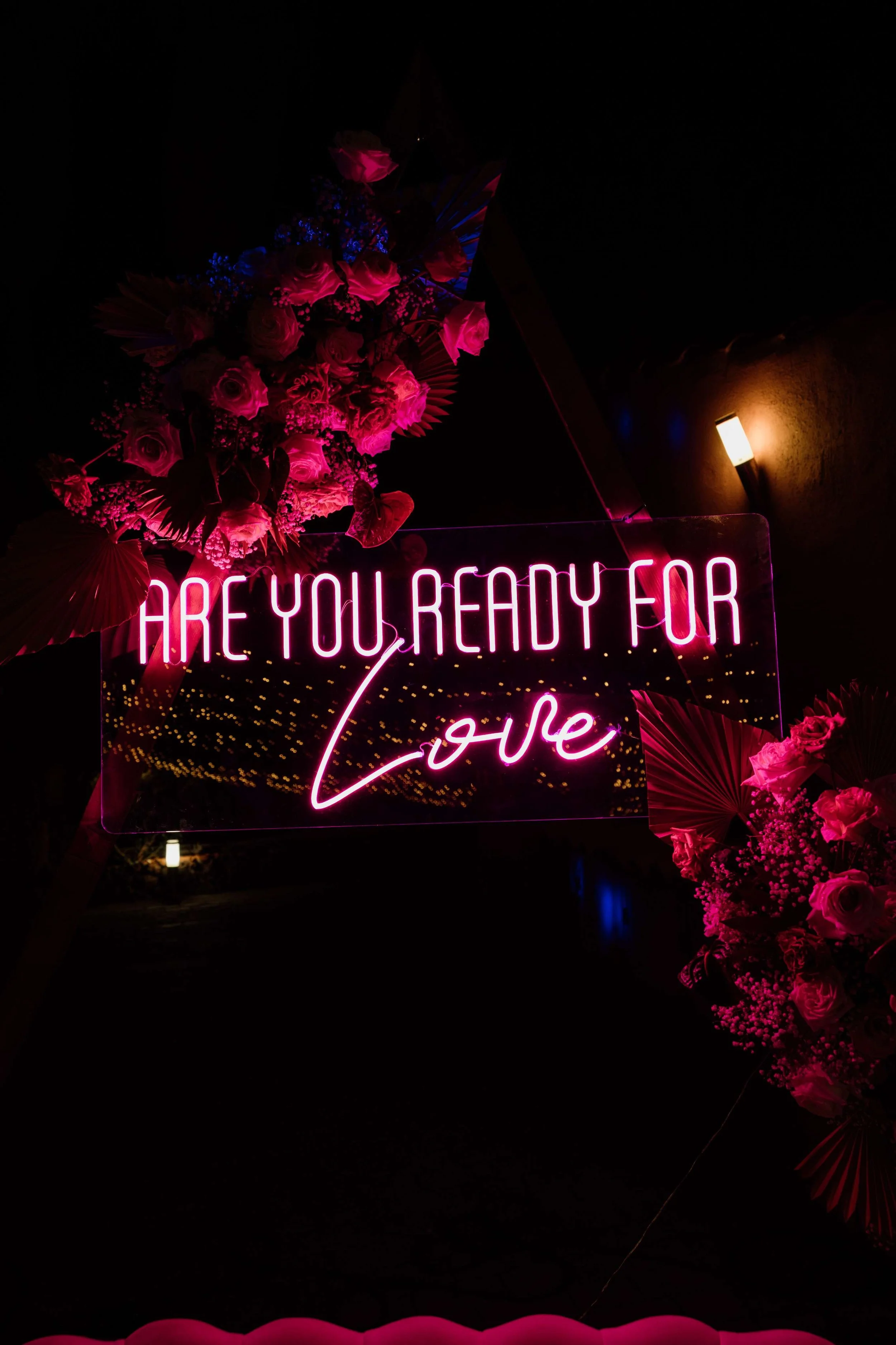 A pink neon sign with the words 'Are you ready for love' surrounded by pink flowers and decorative foliage, set against a dark background.