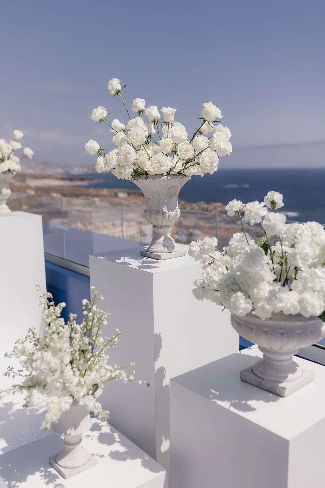 White flower arrangements in elegant vases are displayed on white pedestals outdoors with a view of the ocean and a distant shoreline under a partly cloudy sky.