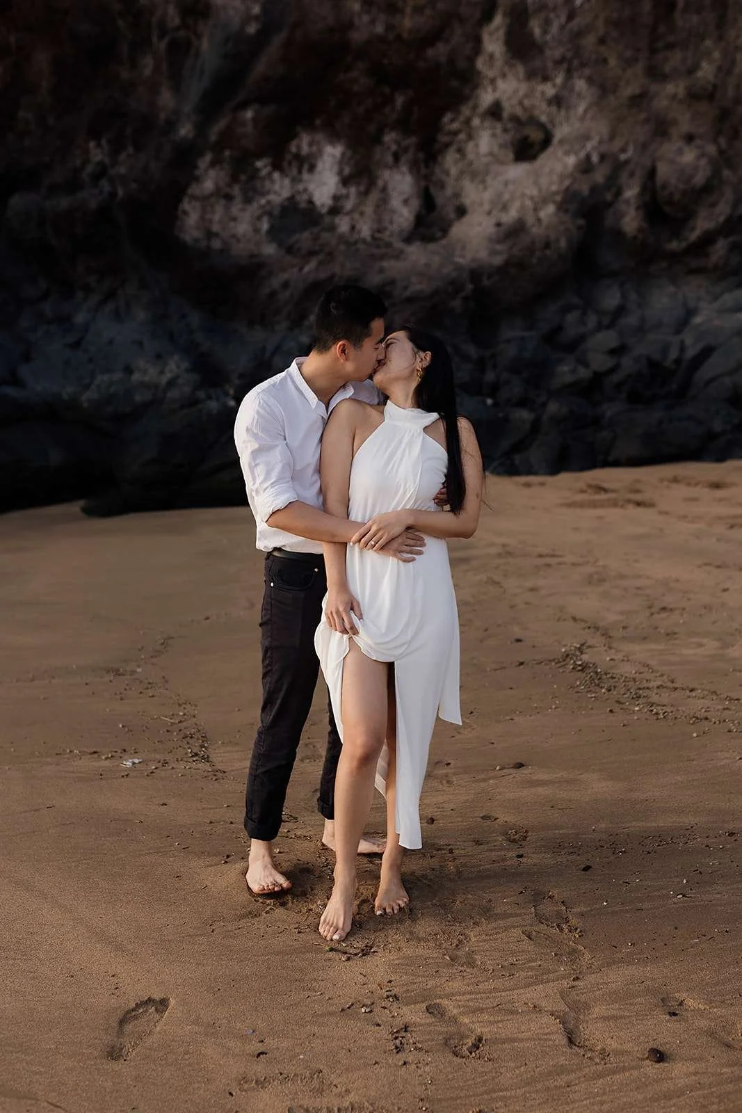 A couple in white and black clothing sharing a kiss while standing barefoot on a sandy beach with a rocky cliff in the background.