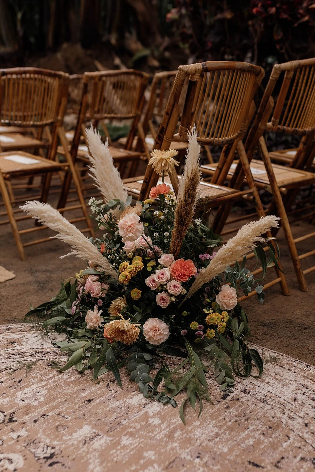 A floral arrangement with pink, peach, and white flowers, including roses and carnations, and tall pampas grass, placed on a woven tablecloth, with wooden chairs in the background.