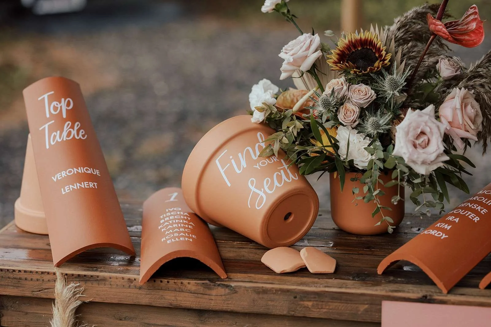 Terracotta flower pots with white and peach roses, sunflower, anthurium, and greenery, some toppled over on a wooden surface with broken pieces nearby, and signs with guest names and table assignments.