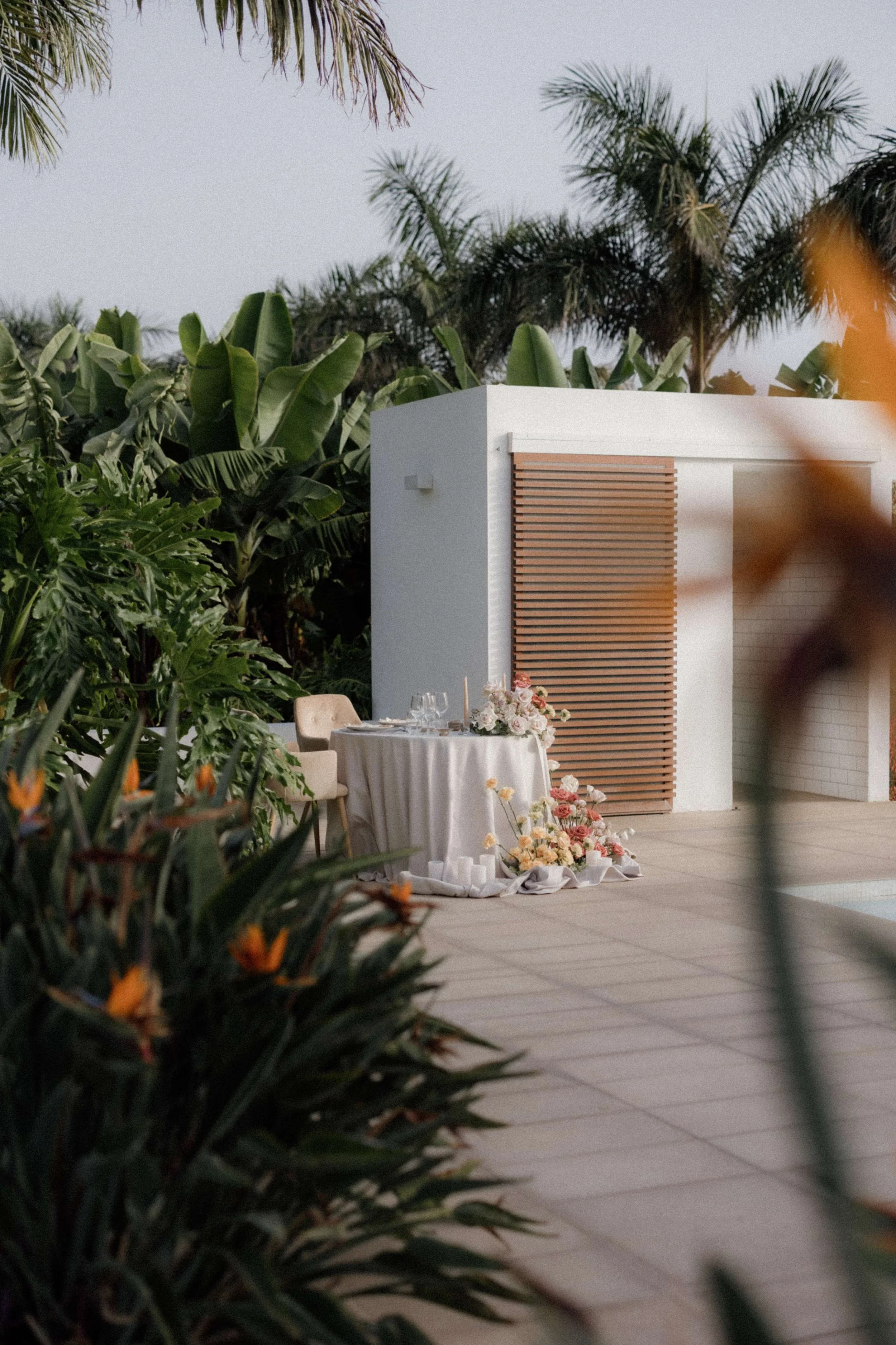 A romantic outdoor dining setup with a round table covered with a white tablecloth, set with wine glasses, plates, and silverware, decorated with a pastel-colored floral centerpiece, situated on a tiled patio surrounded by lush green tropical plants 