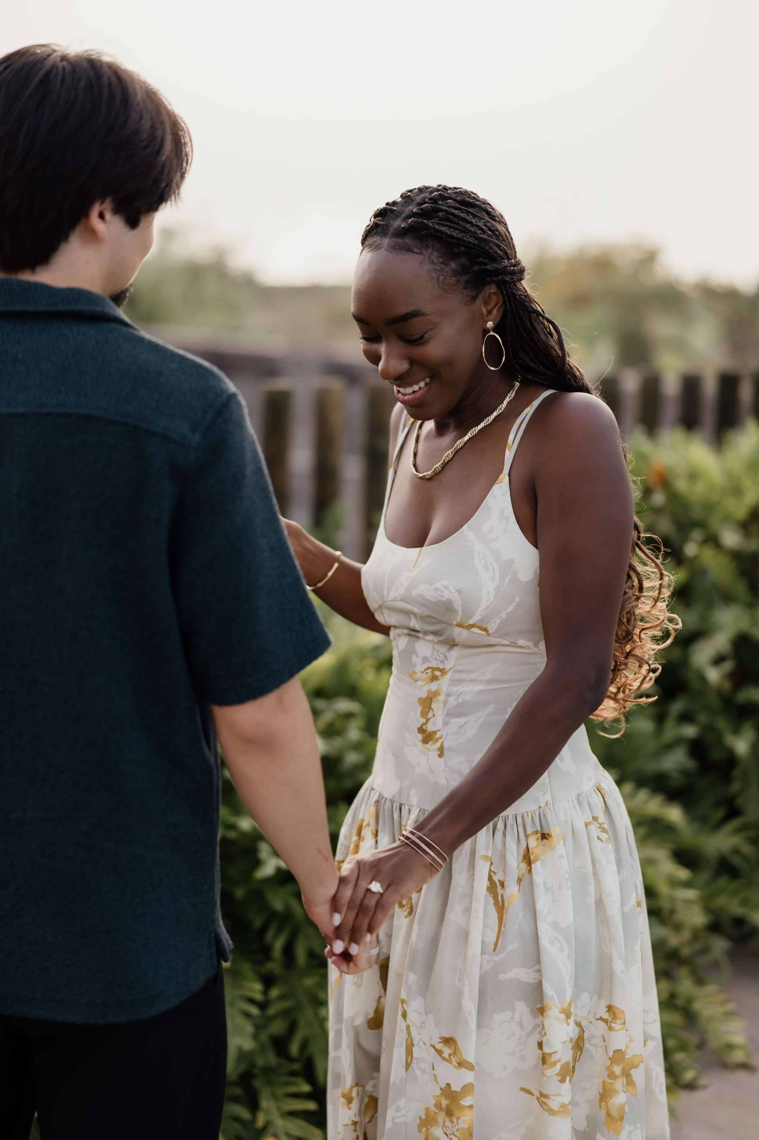 A woman in a cream-colored dress with gold accents holding hands with a man outdoors, smiling with her eyes closed.
