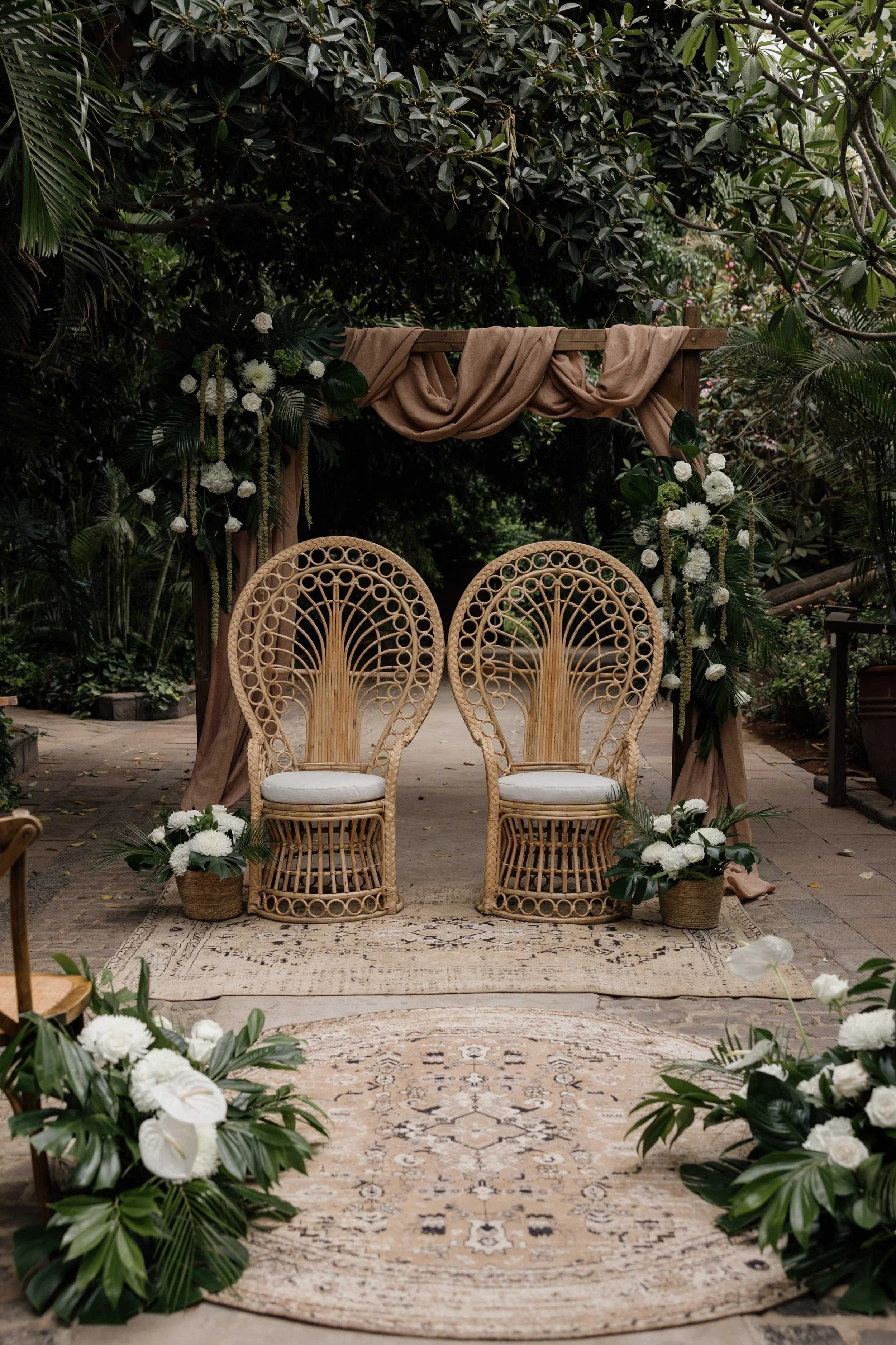 A wedding or special event setup with two wicker peacock chairs in front of a floral arch, decorated with white flowers and greenery, on a patterned rug in an outdoor garden setting.