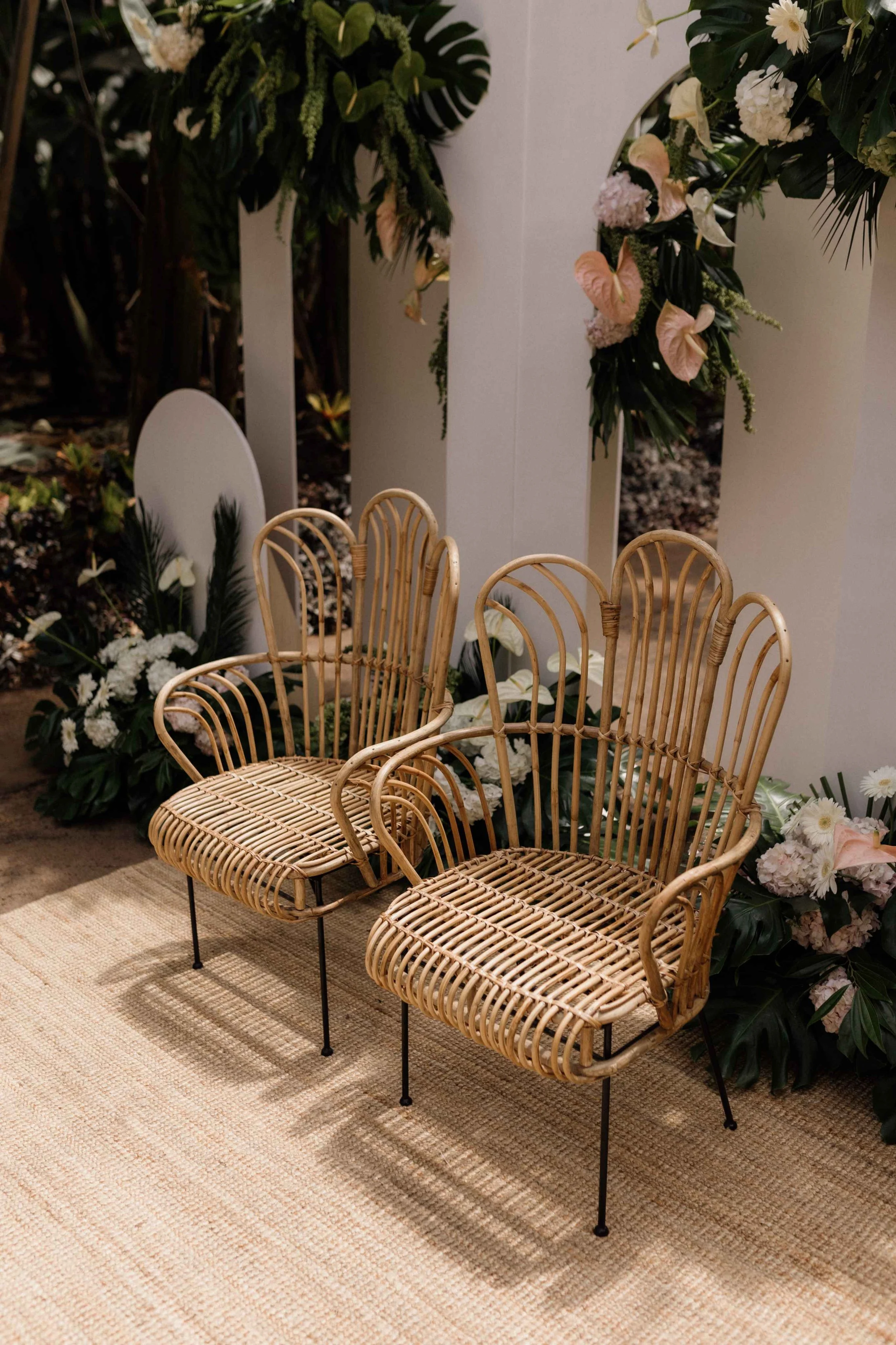 Two rattan chairs with black metal legs placed on a beige rug in front of a floral backdrop, possibly for a wedding or special event.
