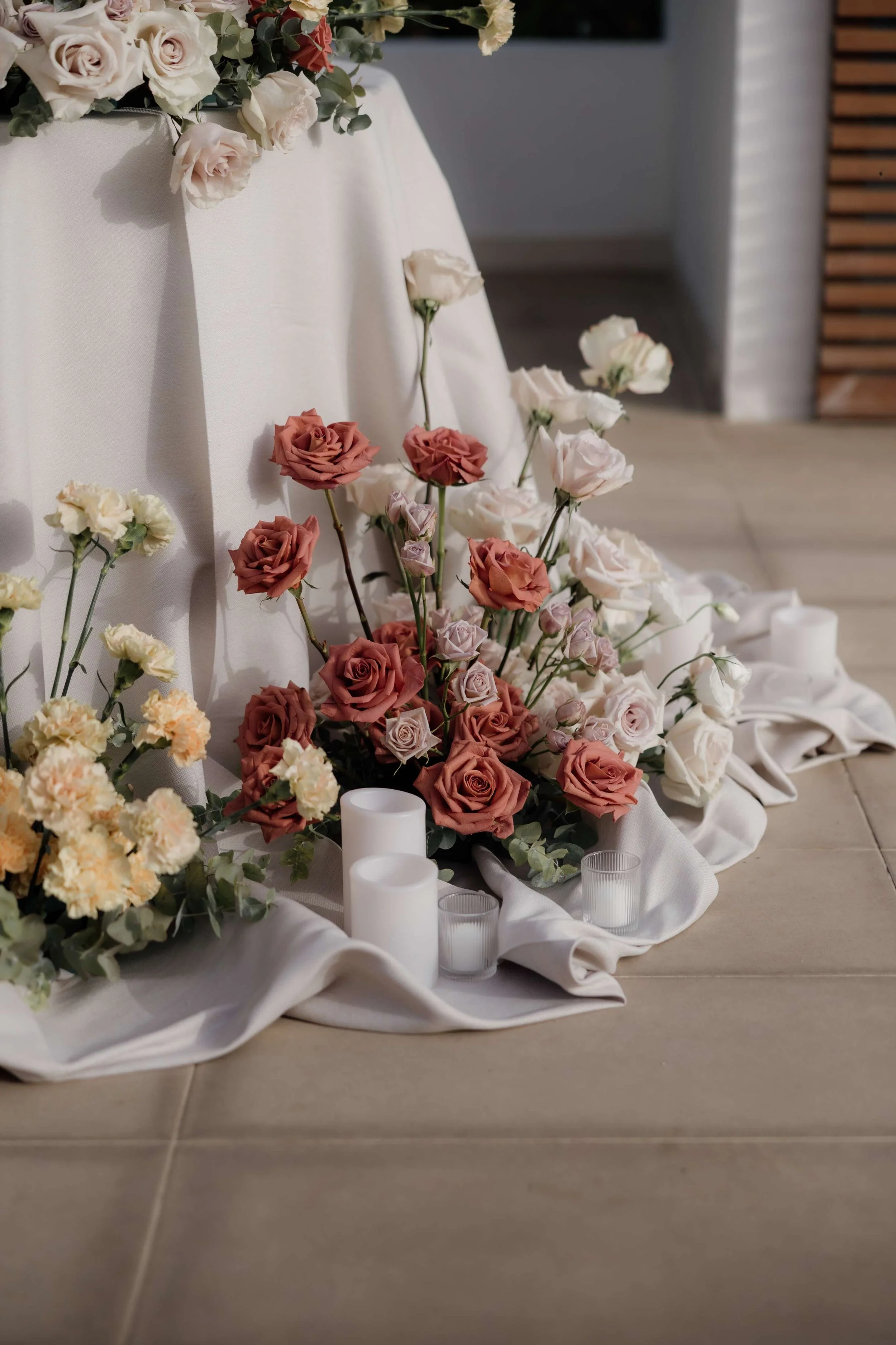 Arrangement of pink, white, and beige roses with candles on a white cloth at a floral display or memorial.