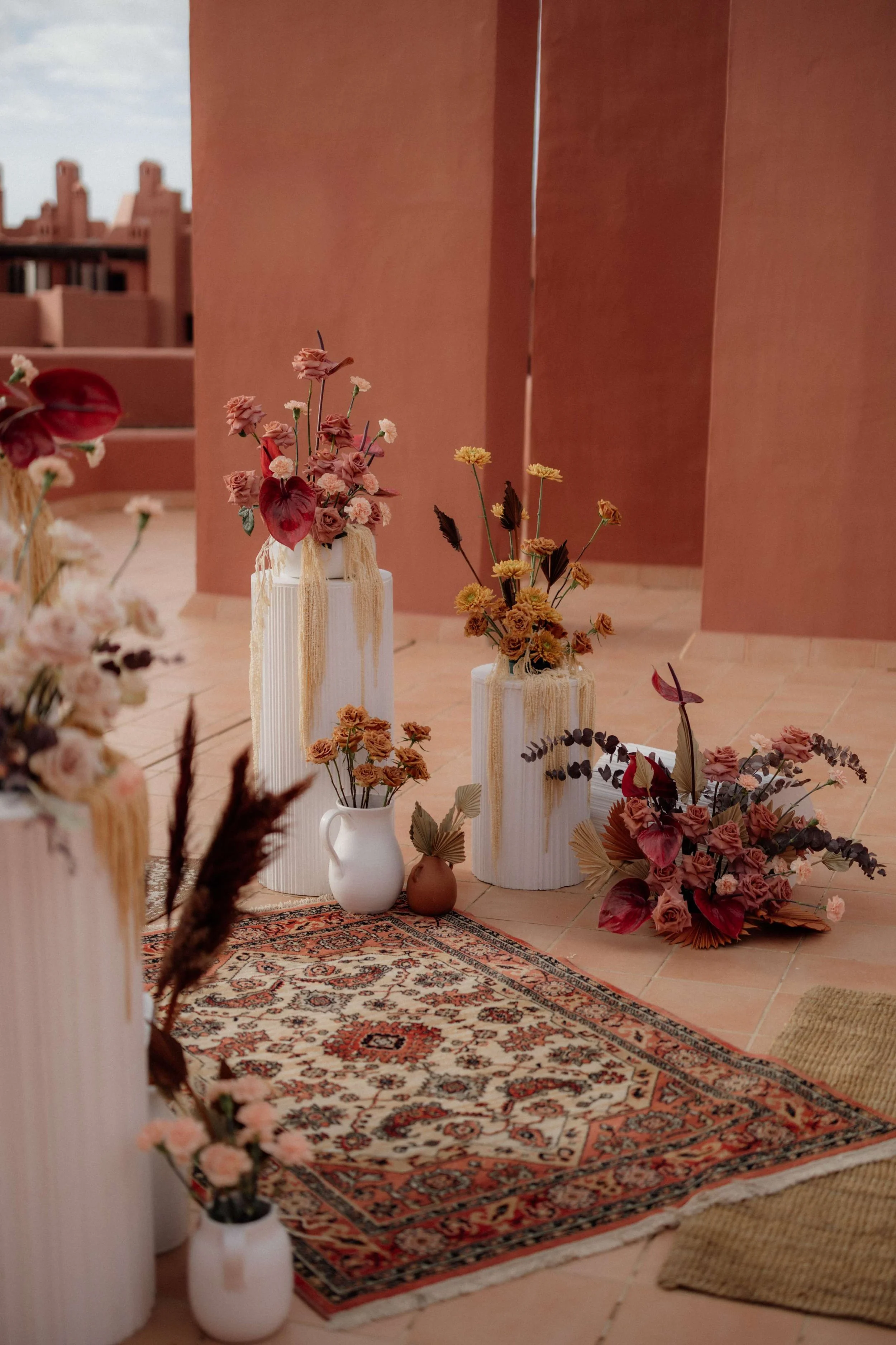 A decorative arrangement of dried flowers in vases arranged on a patterned rug on a terrace with pink walls and cityscape in background.