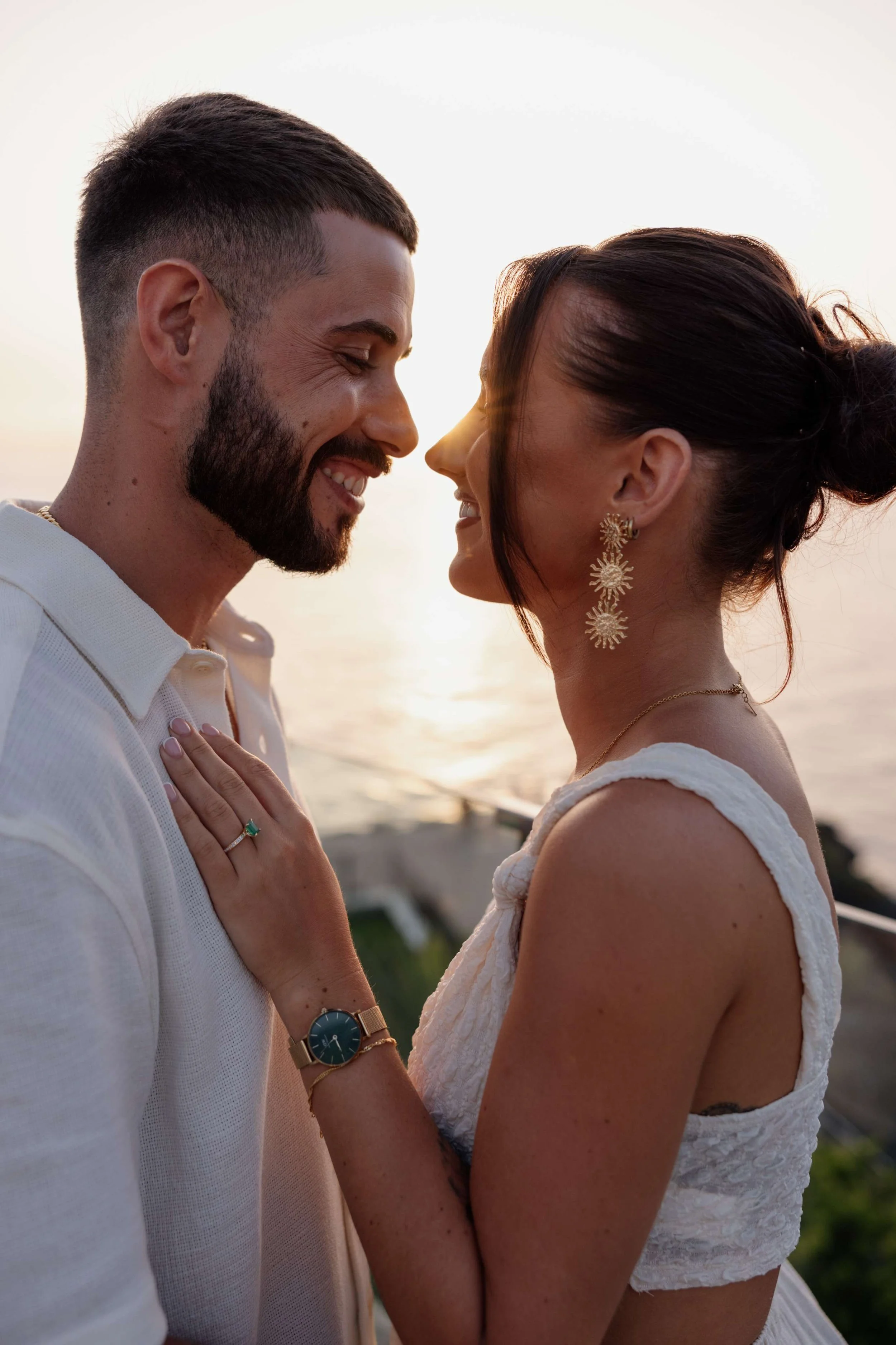 A couple standing close together at sunset, smiling and touching foreheads, by the water. The woman is wearing gold earrings and a sleeveless white top, and the man is wearing a white shirt and a watch.