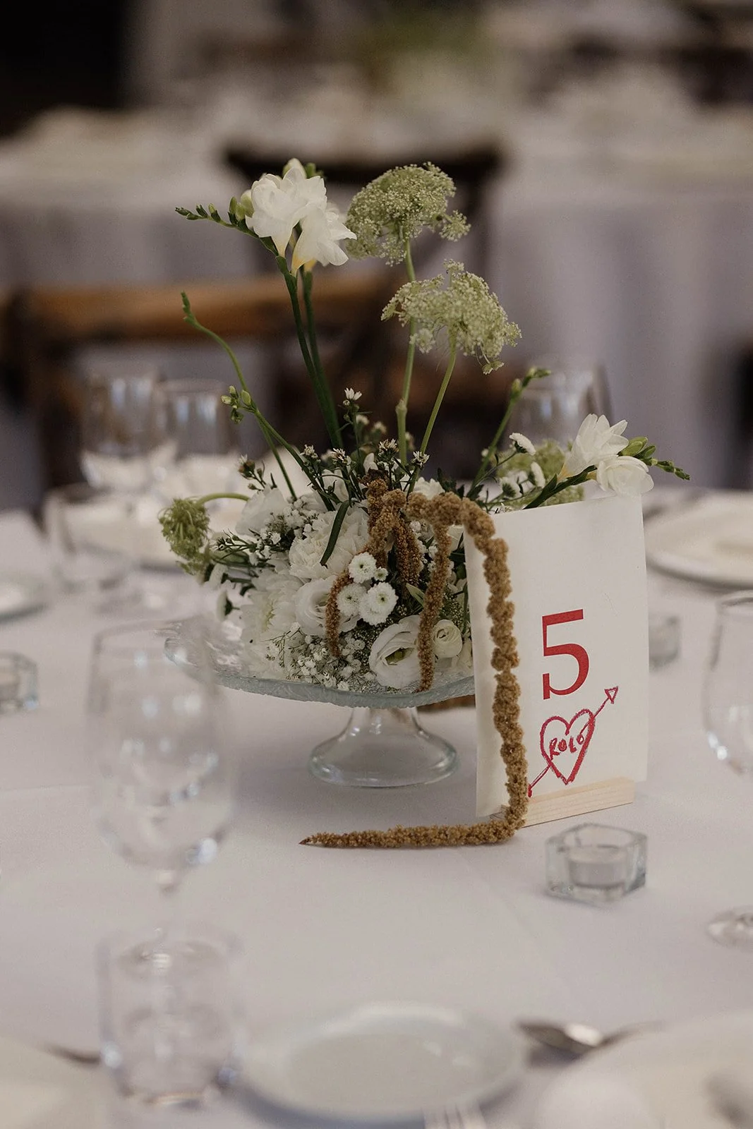 Table centerpiece with white flowers and a table number sign displaying the number 5, with a heart and arrow, and the year 2019, on a white tablecloth surrounded by glassware.
