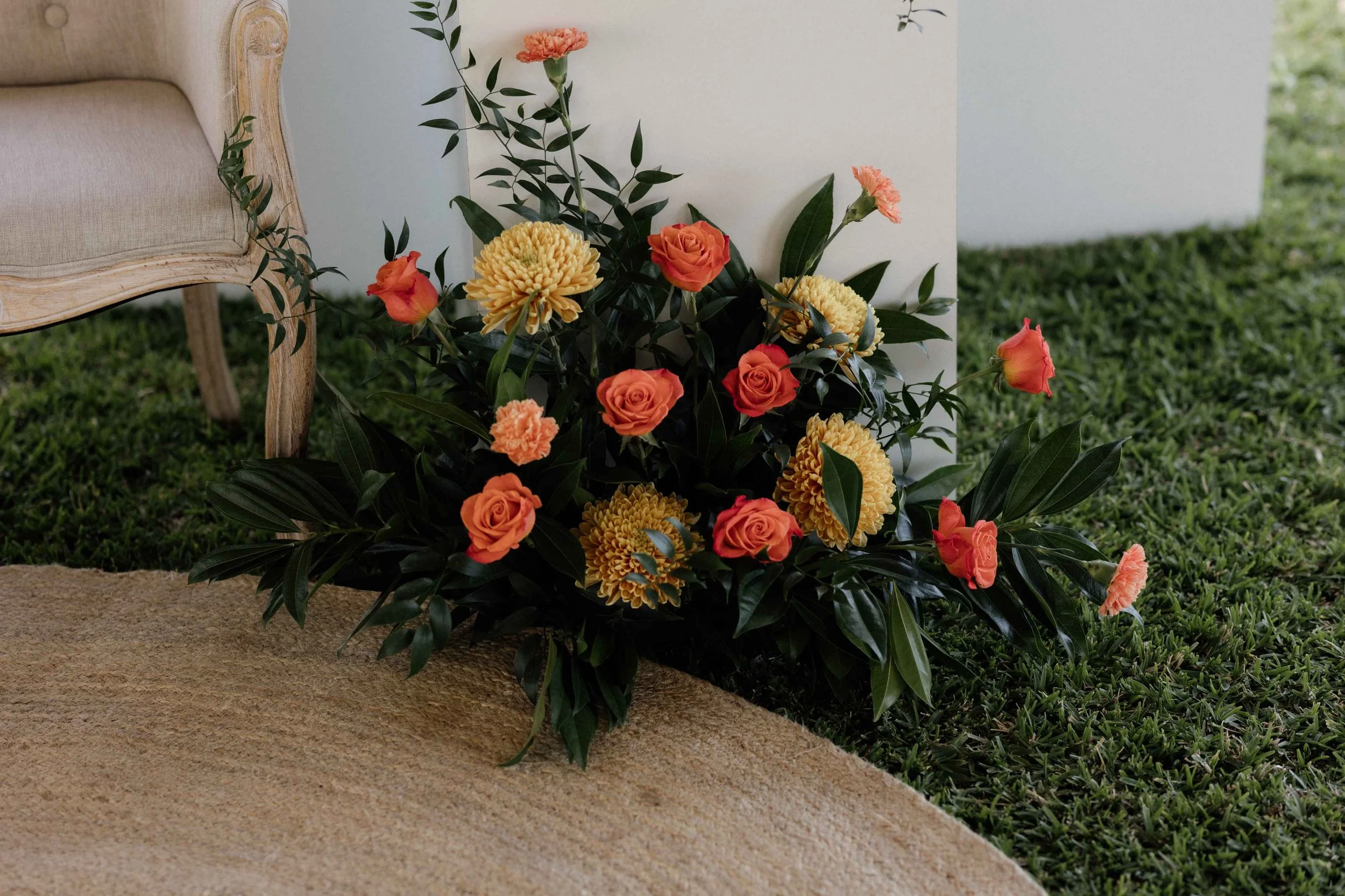 A floral arrangement with peach roses, yellow chrysanthemums, and pink carnations on a grassy area near a beige rug and a light-colored piece of furniture.