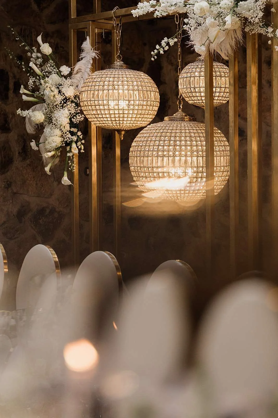 Elegant table setting with two chandeliers hanging from a gold structure, decorated with white flowers and feathers, against a rustic stone wall.