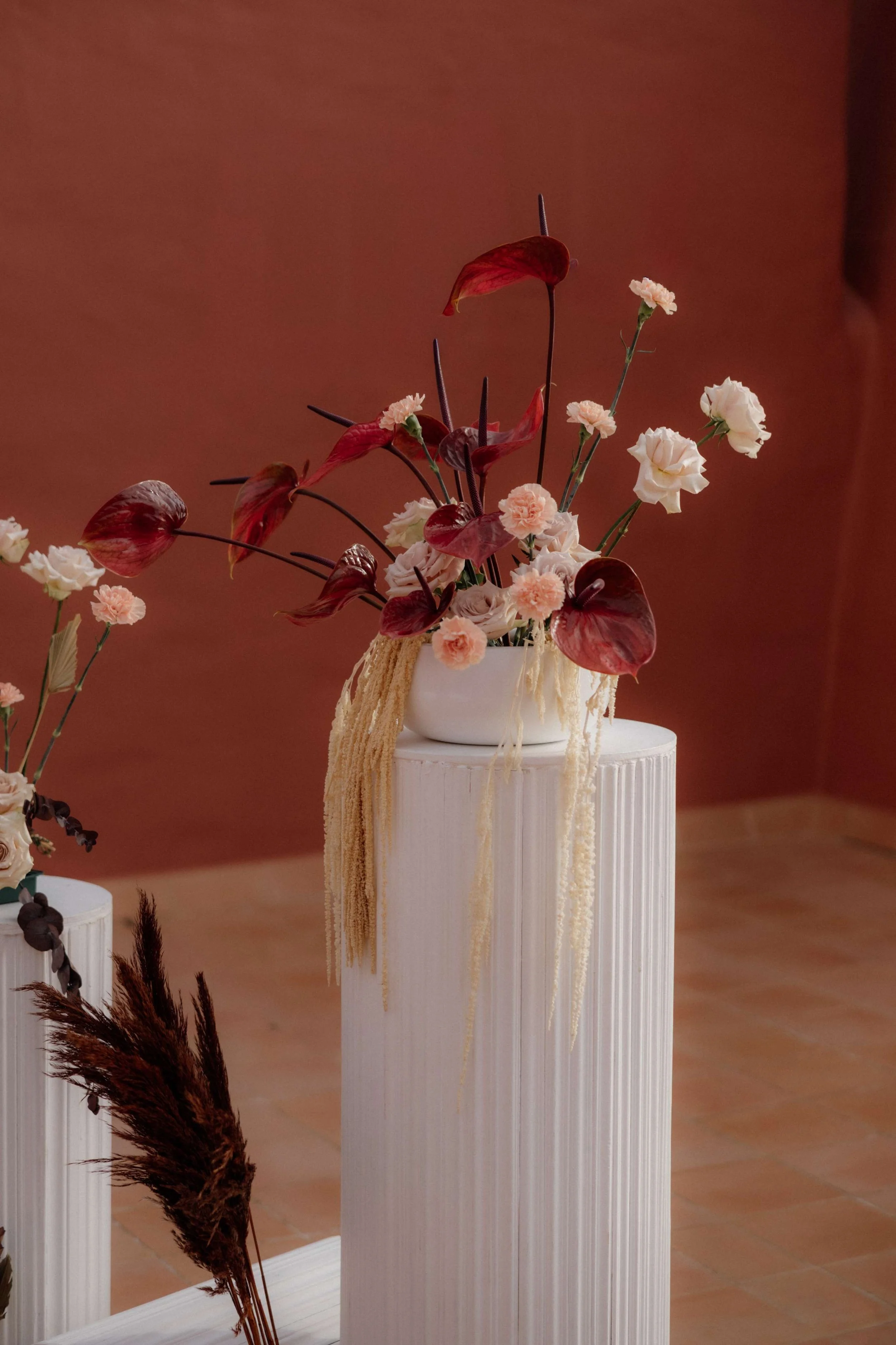 Elegant floral arrangement on a white pedestal, featuring white and pale pink flowers with dark red and purple leaves, against a warm brown background.