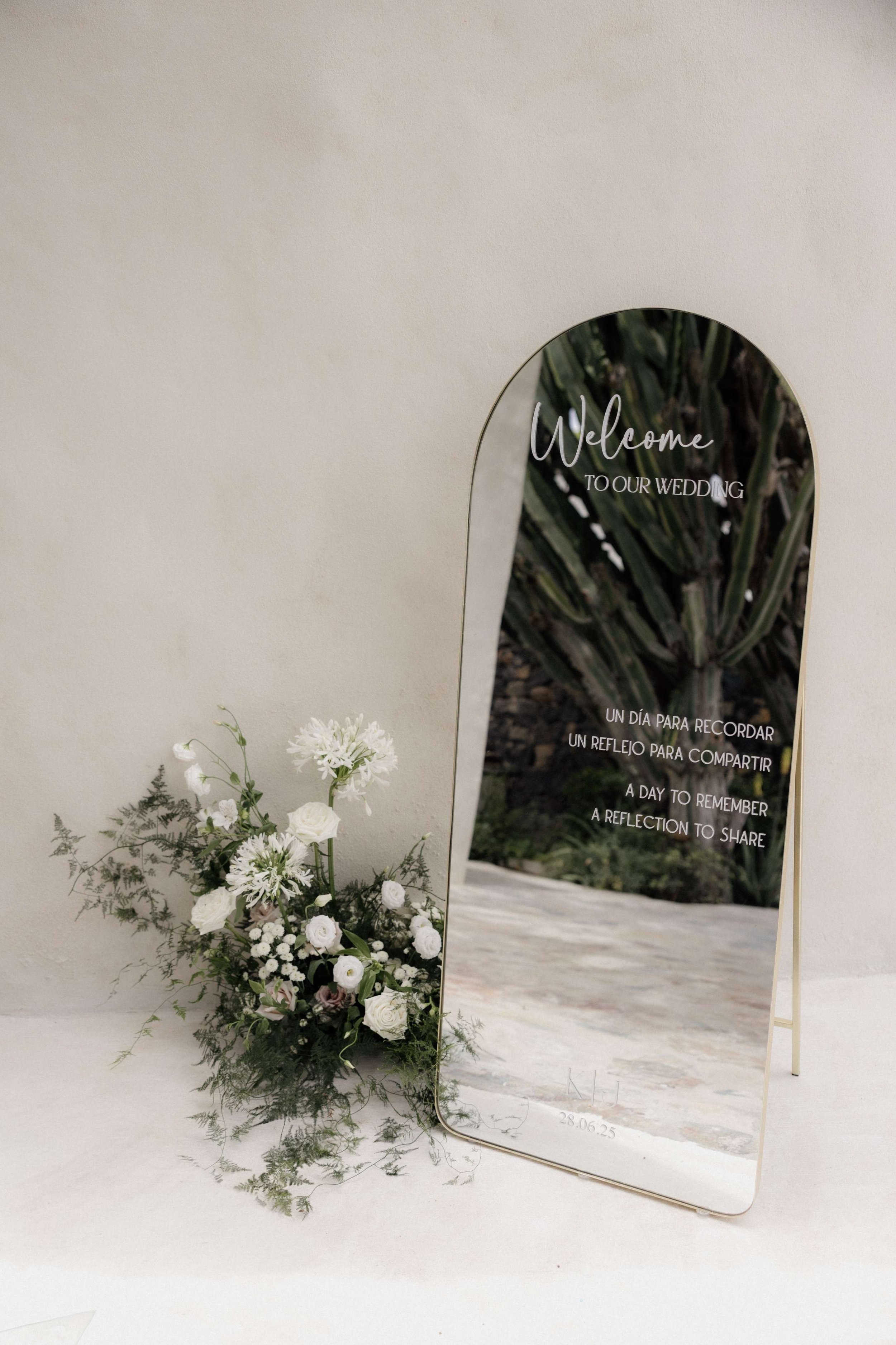 A decorative mirror on a stand with wedding-related text, positioned beside a white floral arrangement on a neutral background.