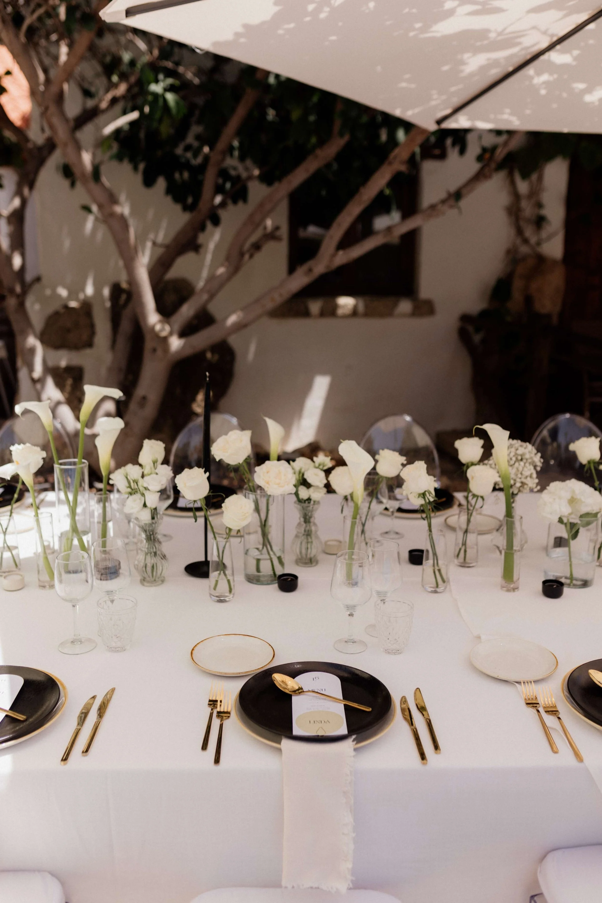 A beautifully set outdoor dining table with white floral arrangements in various glass vases, black and white plates, gold cutlery, and clear glasses under a large white umbrella.