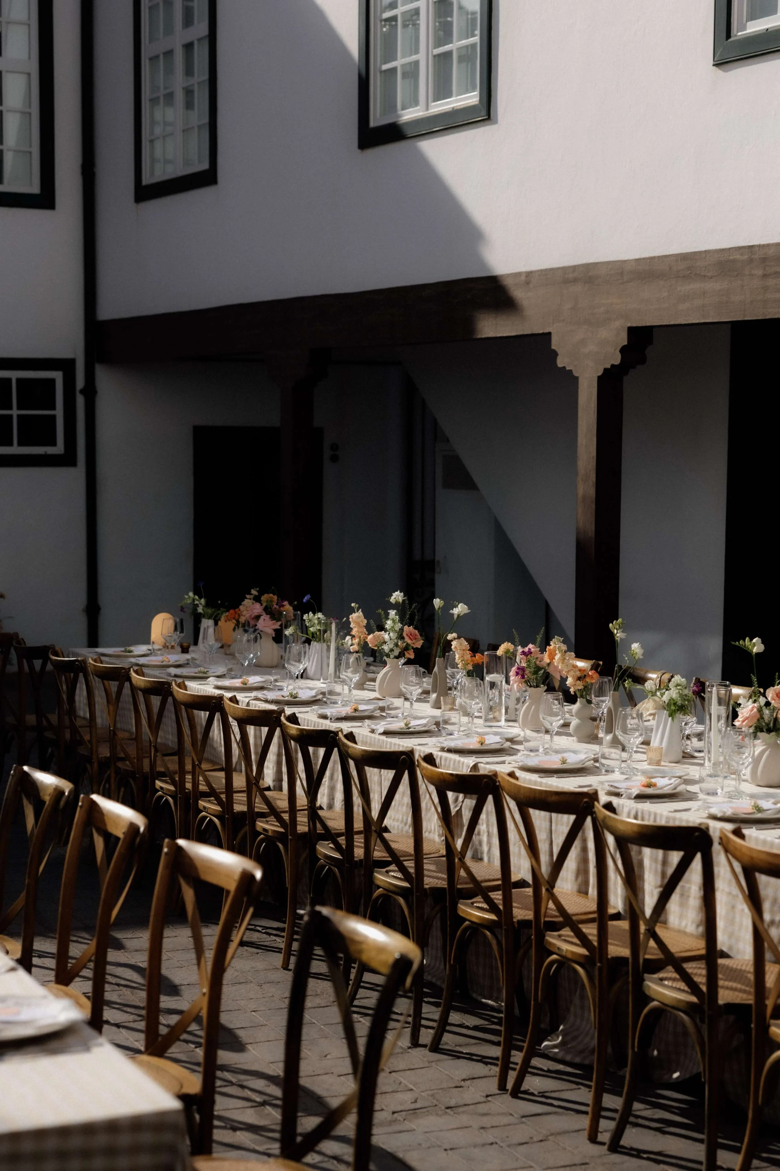 Long outdoor dining table set with plates, silverware, glasses, and pink and white floral centerpieces, with wooden chairs on a brick courtyard next to a white building.