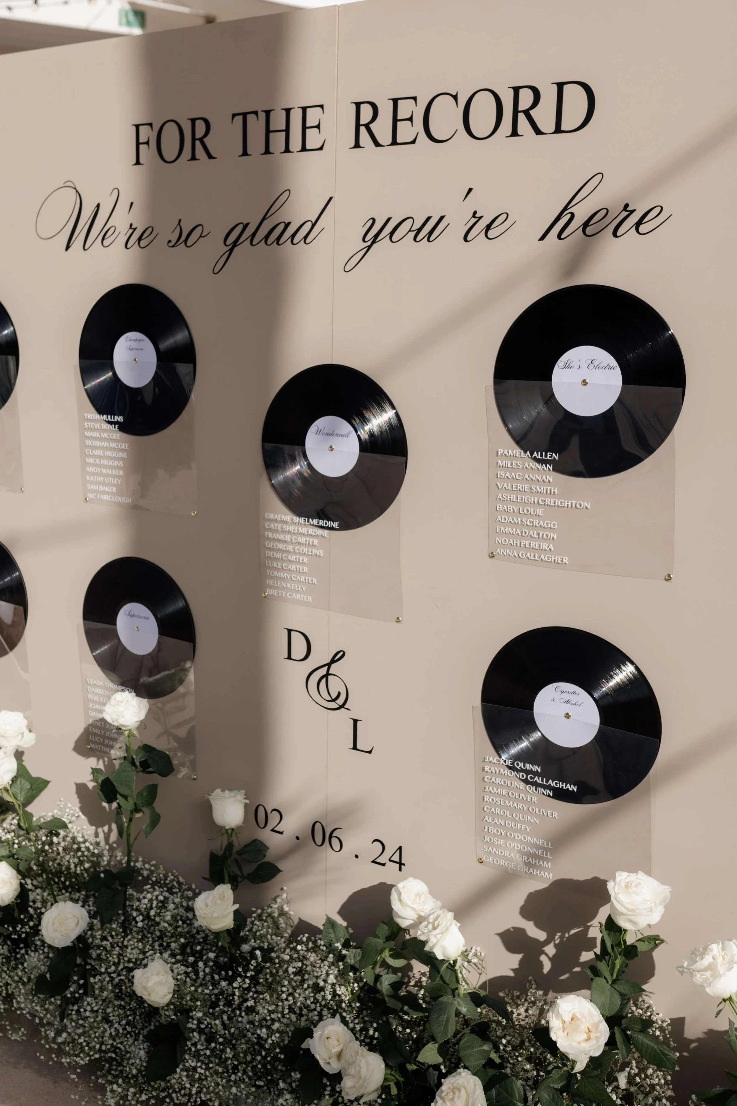 Decorative wall display with vinyl records and guests’ names, celebrating a wedding or special event, with floral arrangements at the base, and the phrase 'For the Record' and 'We're so glad you're here' written on the wall.