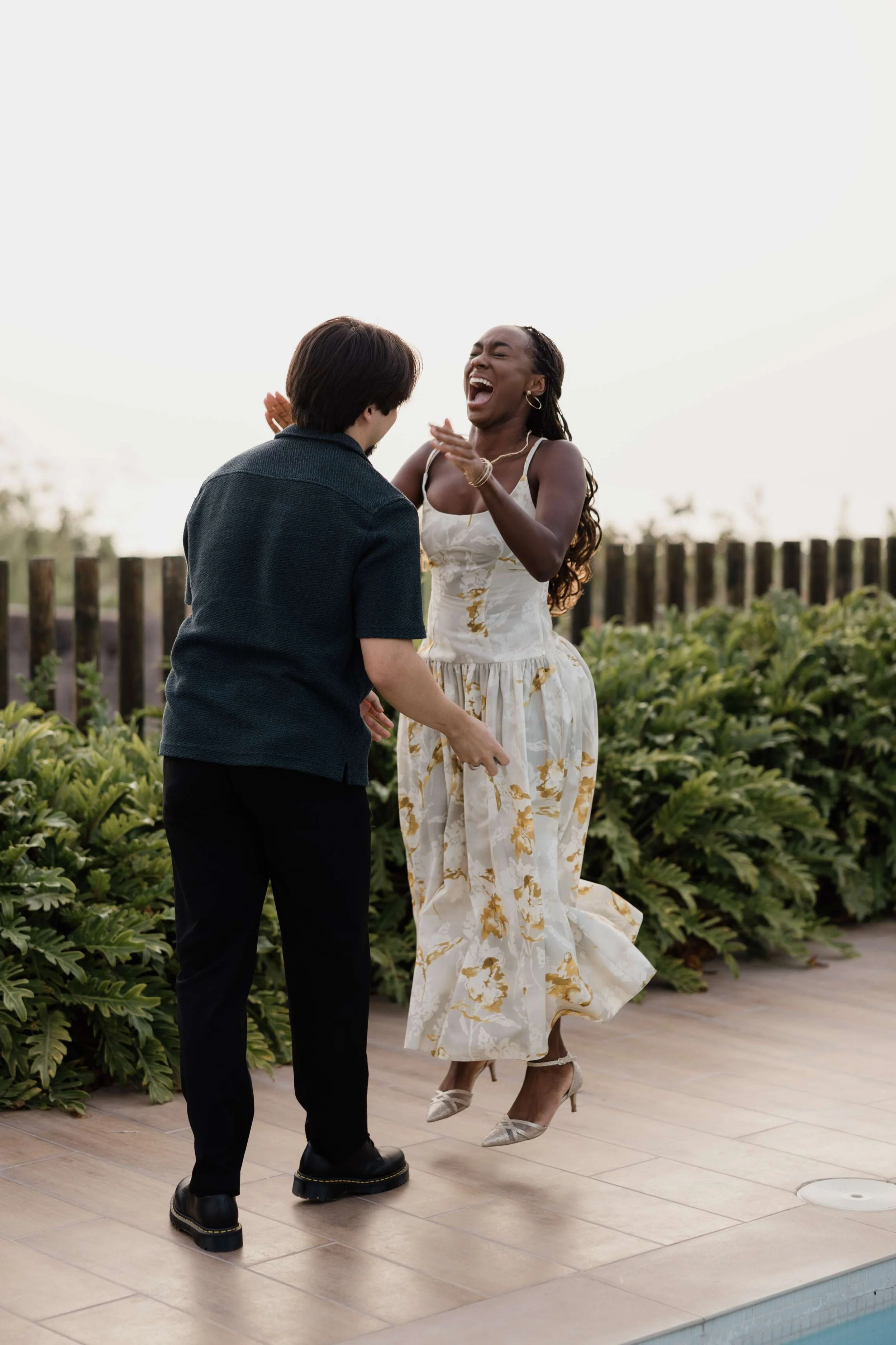 A woman in a white floral dress and high heels is joyfully dancing with a man in dark clothing outdoors near a pool, with green plants and a wooden fence in the background.