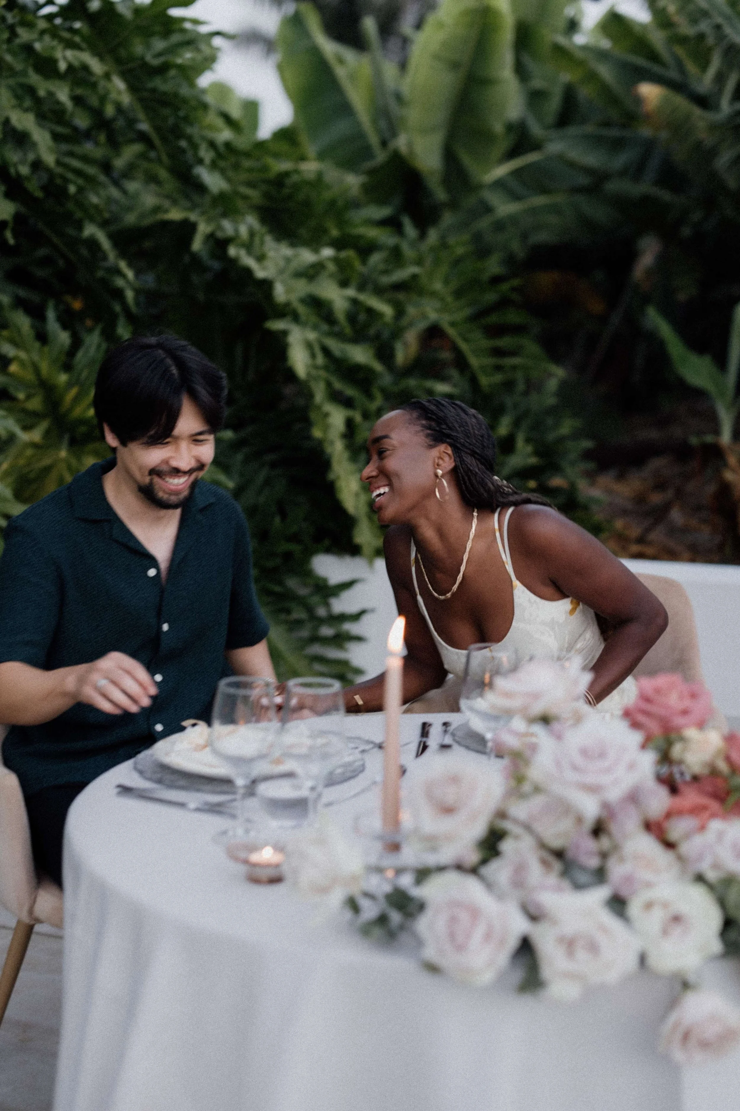 A man and woman laughing at a table with pink and white flowers, candles, and dinnerware, during a dinner celebration.