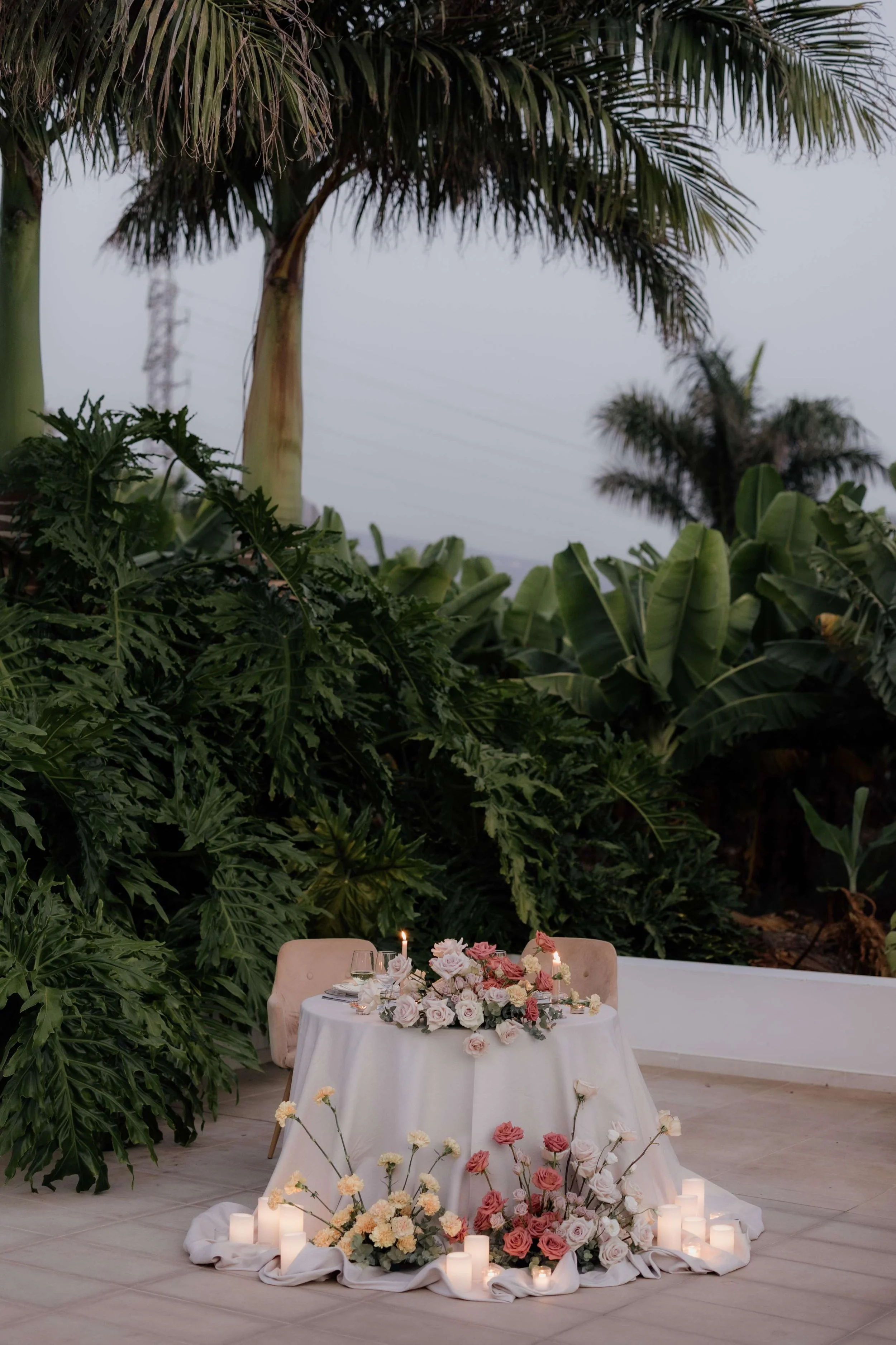A table decorated with pink and white flowers, surrounded by candles, set in an outdoor area amid lush green tropical plants and trees.