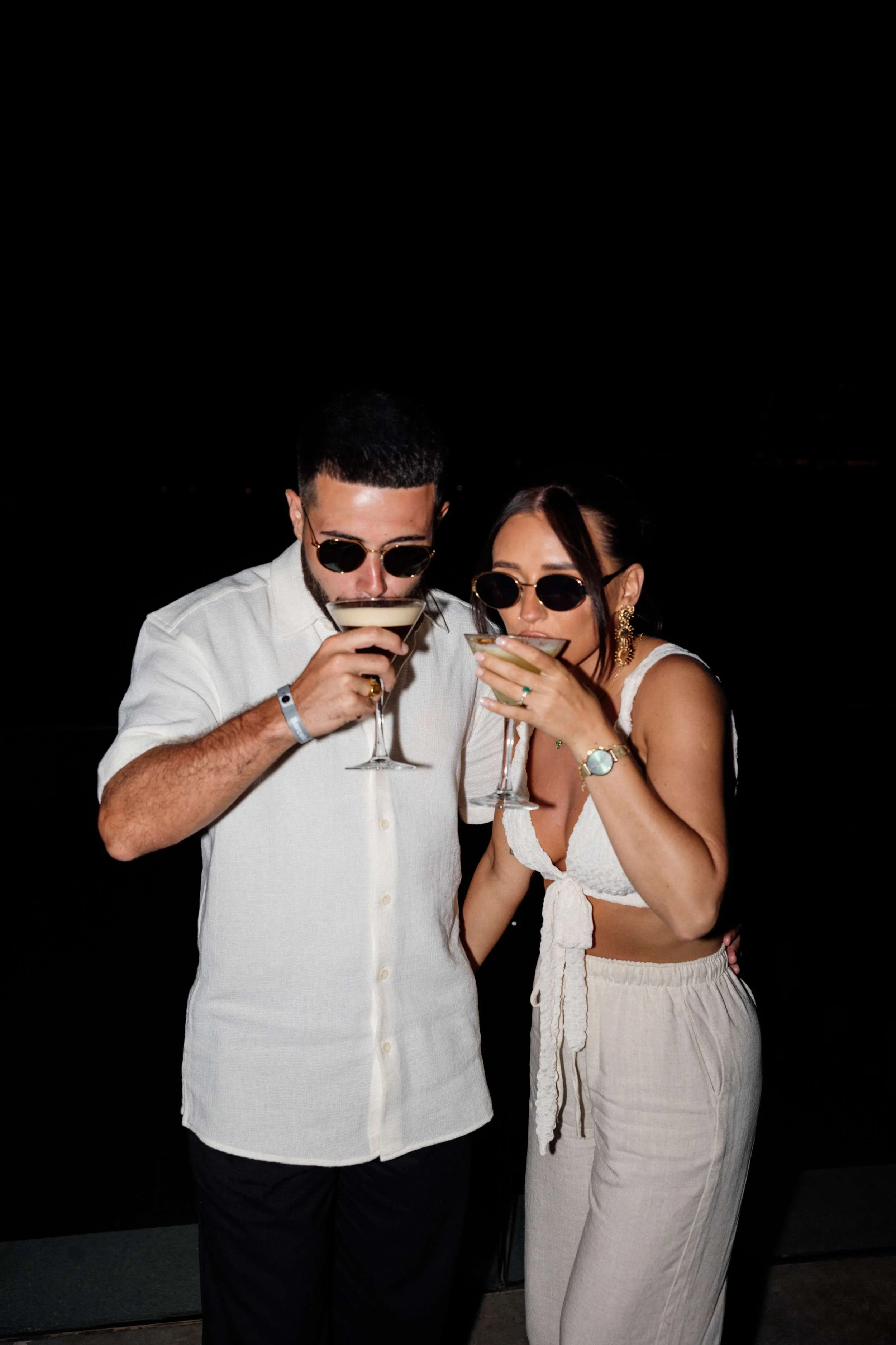 Couple in white clothing and sunglasses holding cocktails at night.