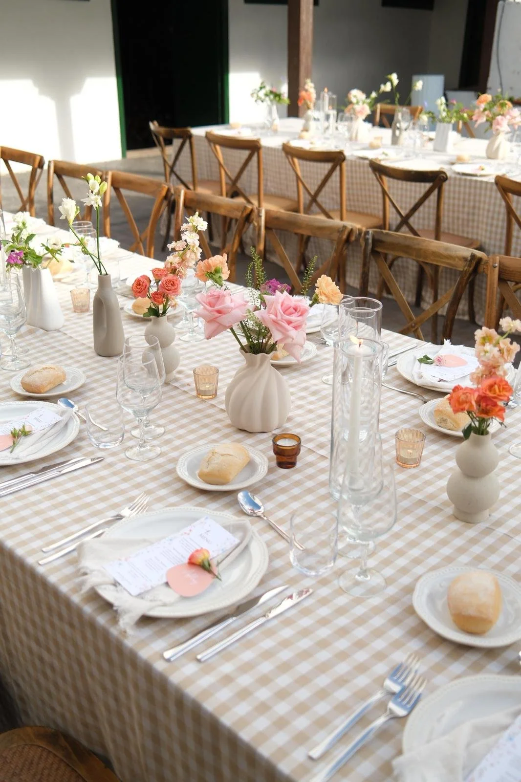 A elegantly set dining table with floral centerpieces, candles, and place settings with bread rolls in a bright, inviting space.