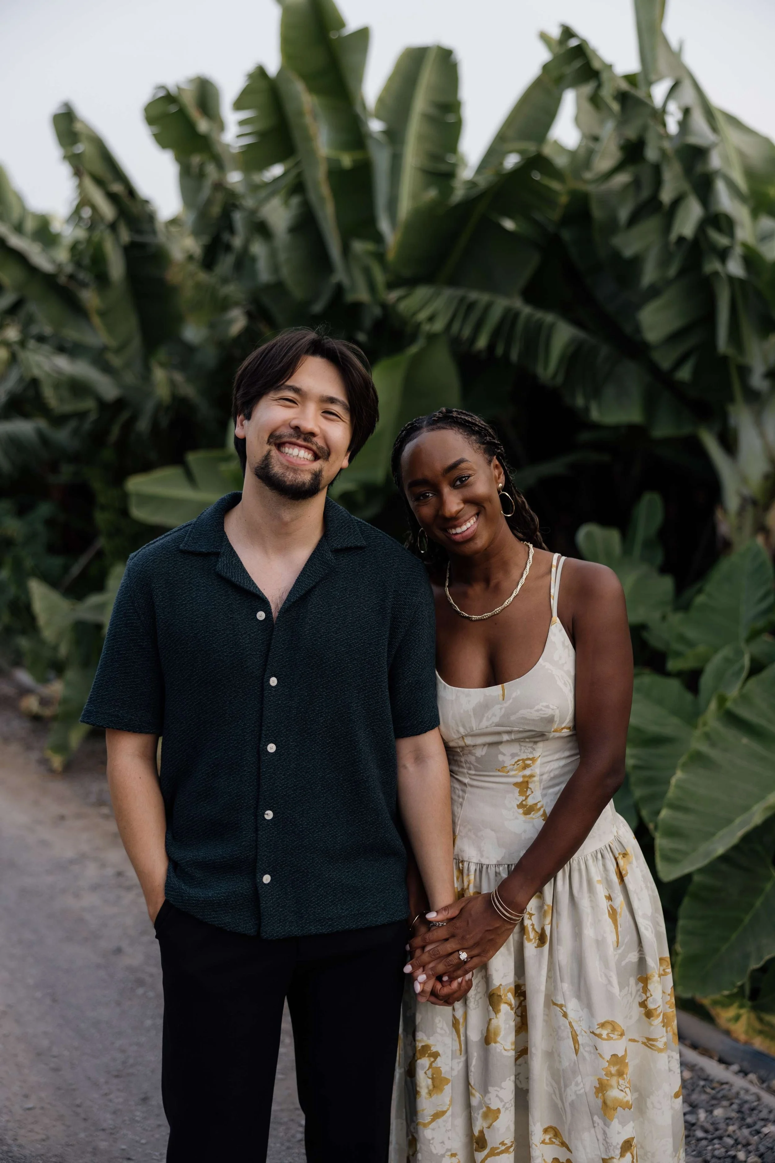 A smiling couple holding hands in front of large leafy green plants, likely banana trees, outdoors during daytime.