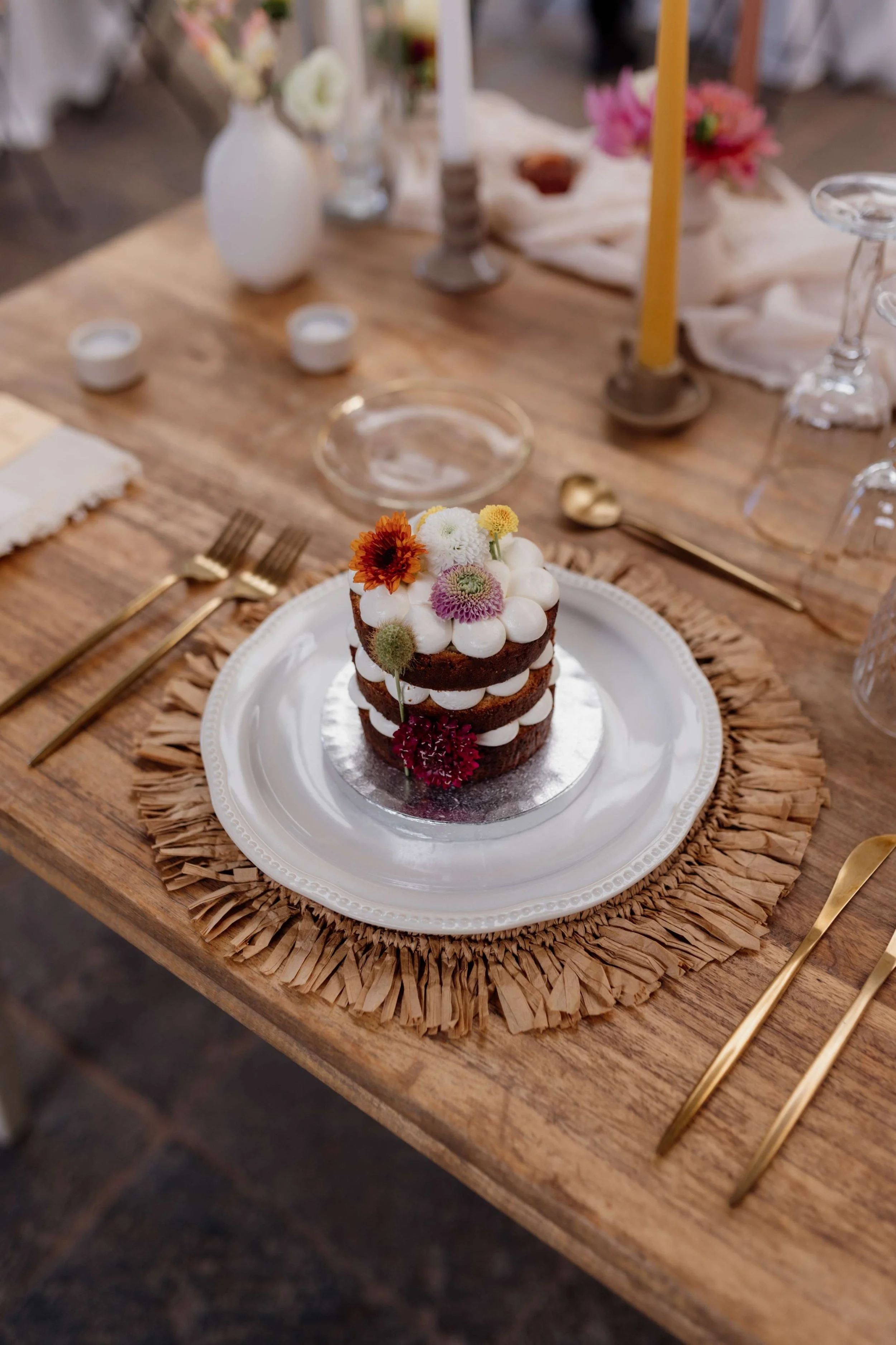 Decorated three-layer chocolate cake with white frosting and edible flowers on a white plate, placed on a rustic wooden table at a celebration or wedding event.