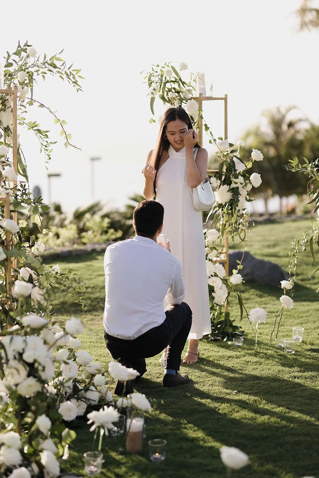 A man proposing to a woman at an outdoor wedding arch decorated with white flowers and greenery, during sunset.