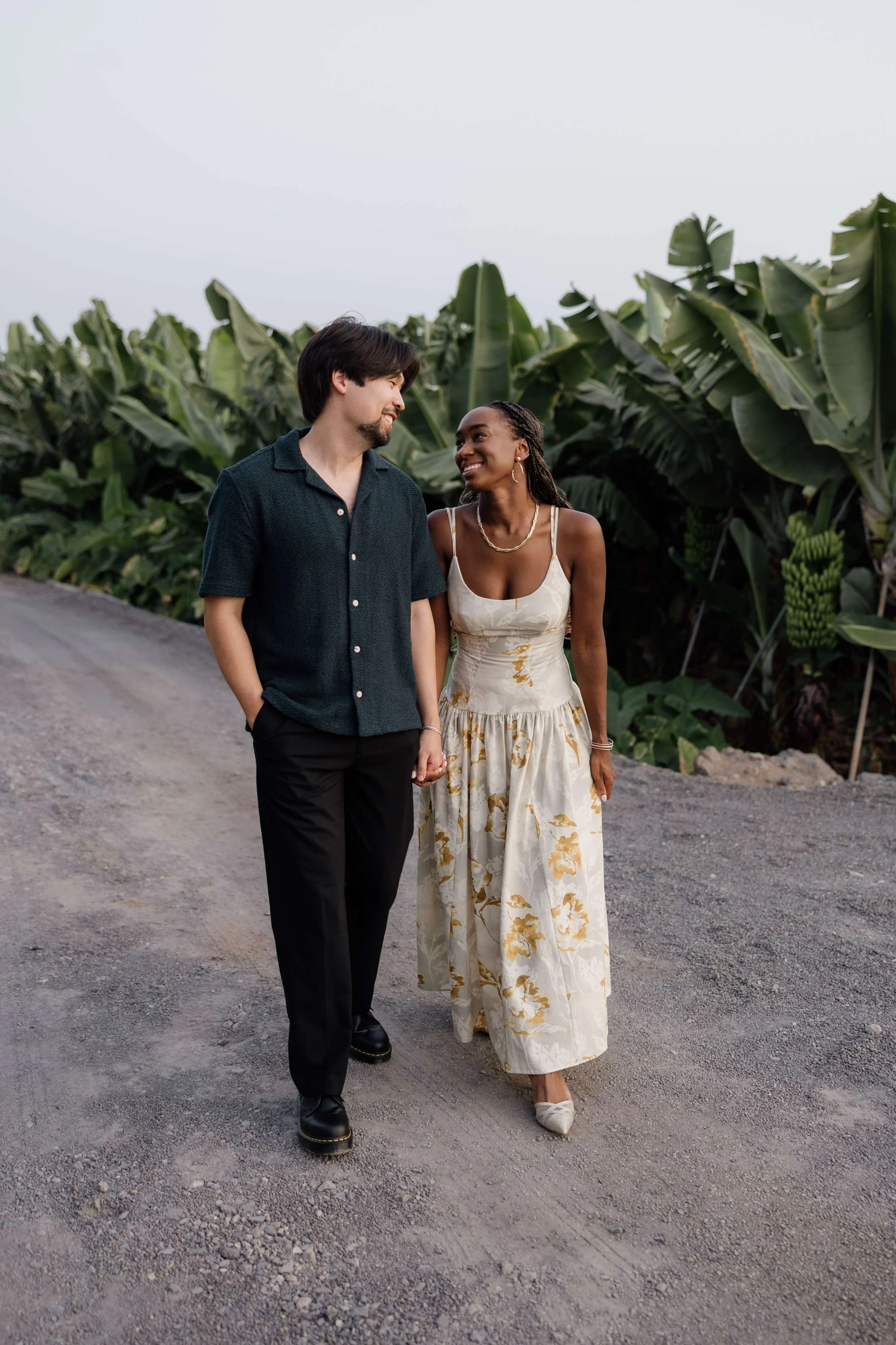 A couple walking hand in hand on a dirt path, smiling at each other, with banana plants in the background, during the daytime.