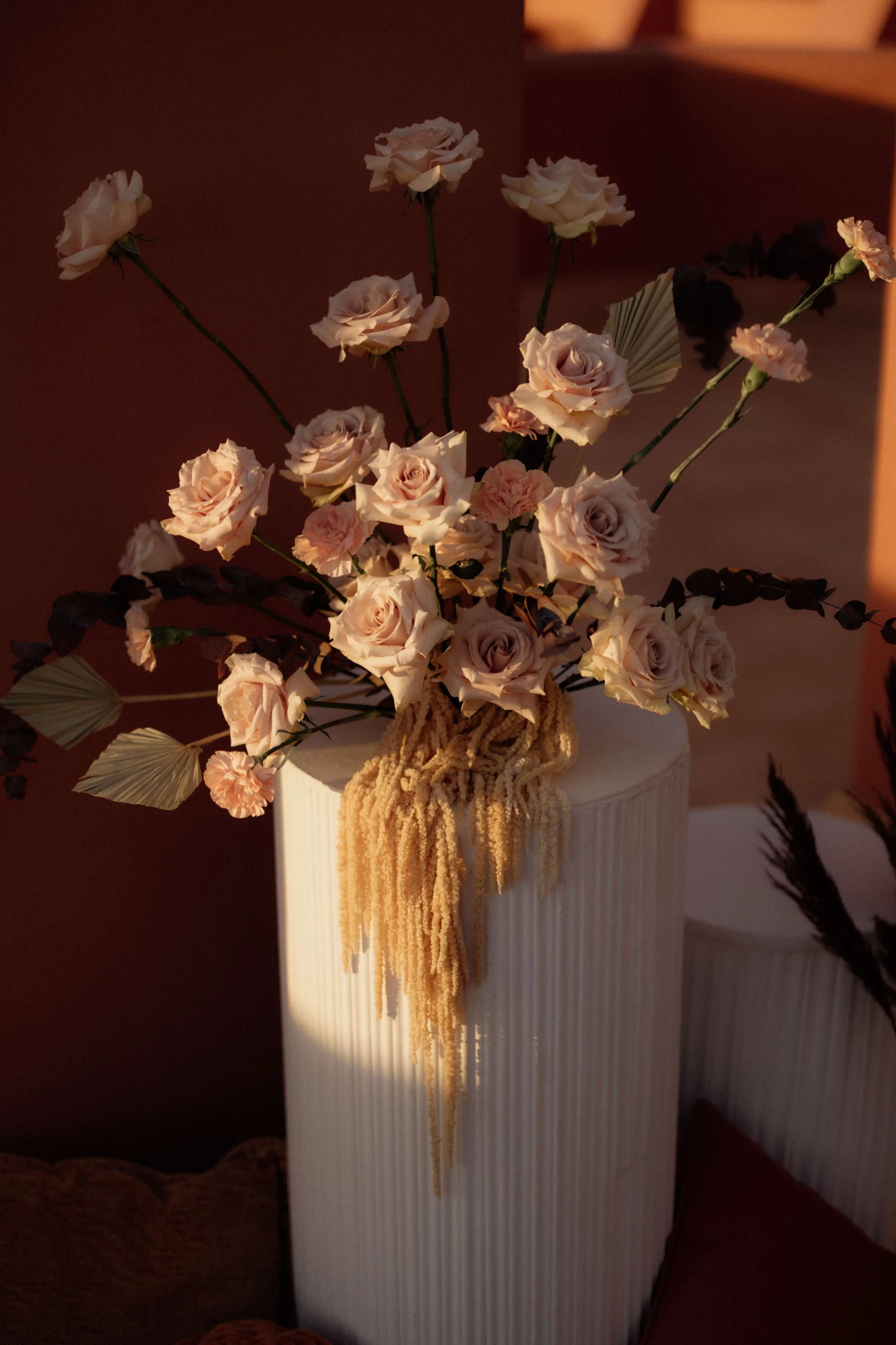 A tall cream-colored vase with vertical ridges holding a bouquet of pale pink roses, pink carnations, and dark purple leaves, with some dried beige hanging embellishments, against a warm-toned background.
