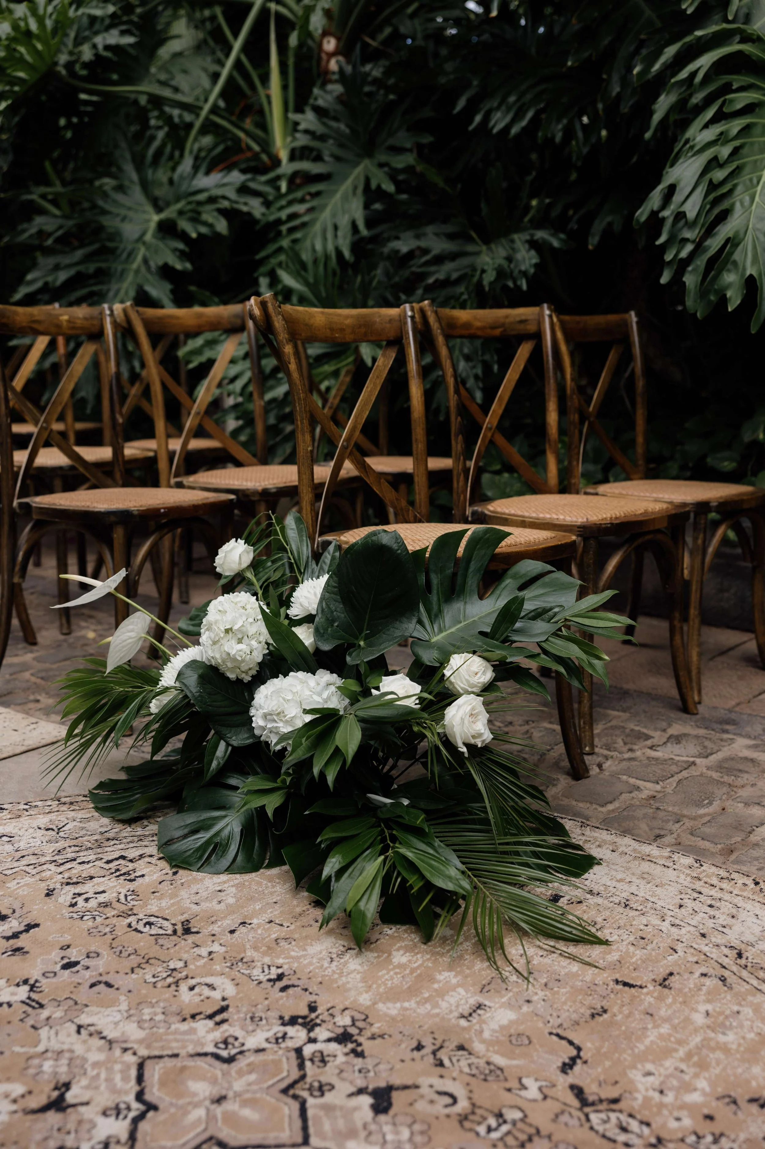 A floral arrangement with white flowers and green tropical leaves on a vintage rug, set in front of wooden chairs in a lush indoor garden.