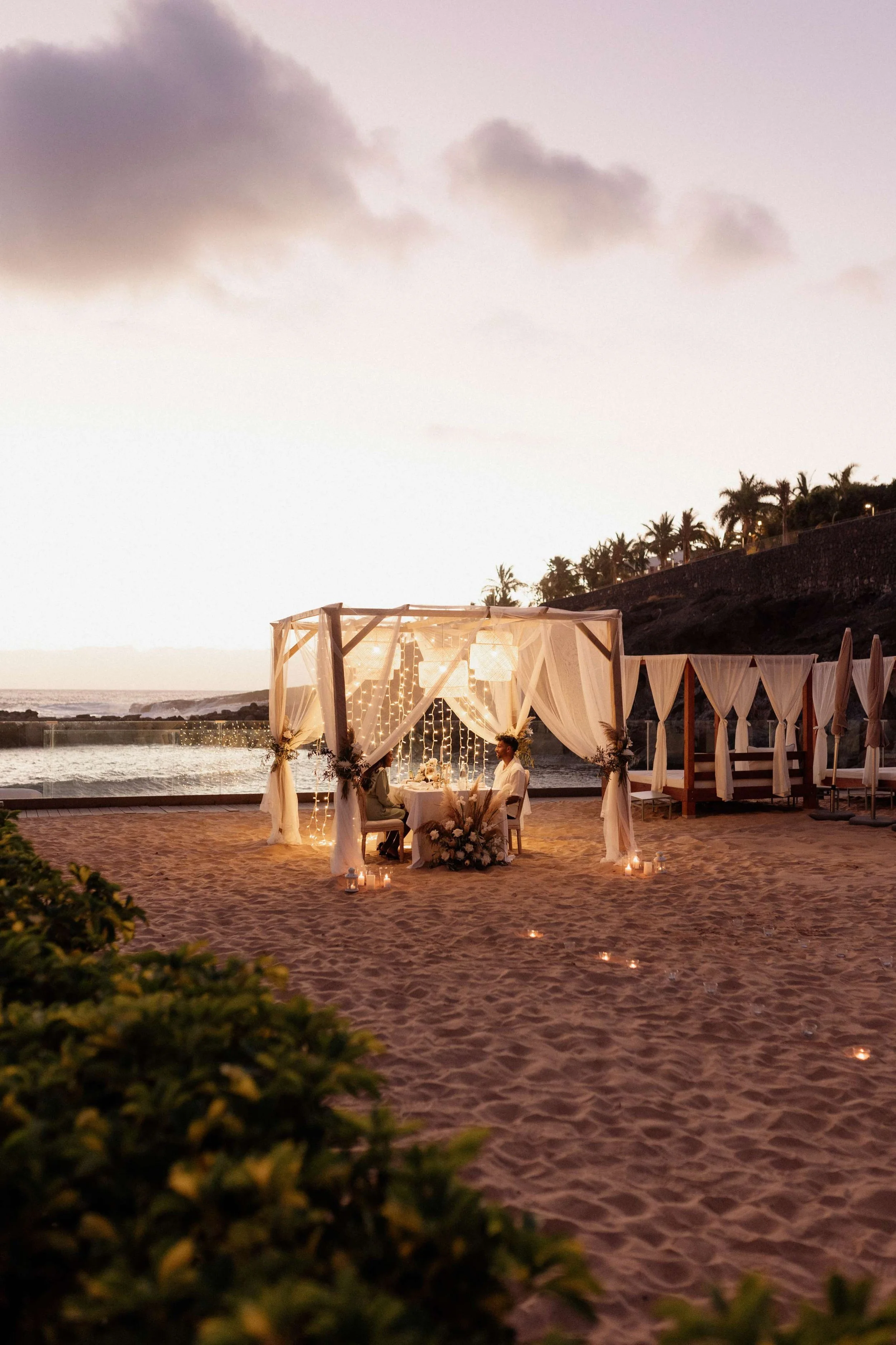 A beach wedding ceremony setup with a canopy decorated with white drapes and string lights at sunset, a couple seated at a table, candles on the sand, and palm trees in the background.