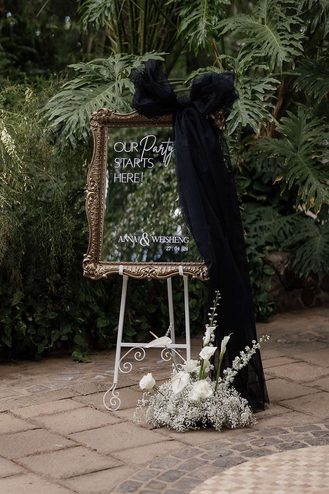 A decorative sign with a black ribbon and flowers in front, indicating a celebration, possibly a wedding, in an outdoor setting surrounded by green foliage.