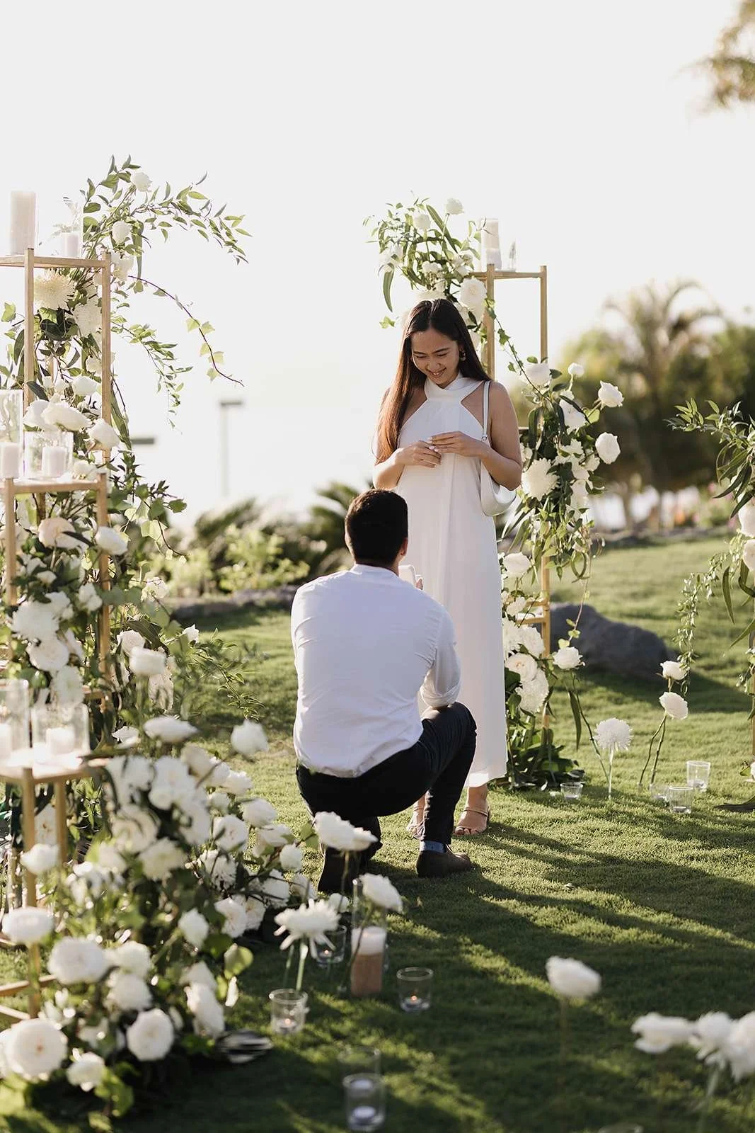 A couple shares a romantic moment outdoors during a wedding ceremony, with the man kneeling and the woman smiling, surrounded by white flowers and greenery in a garden setting.