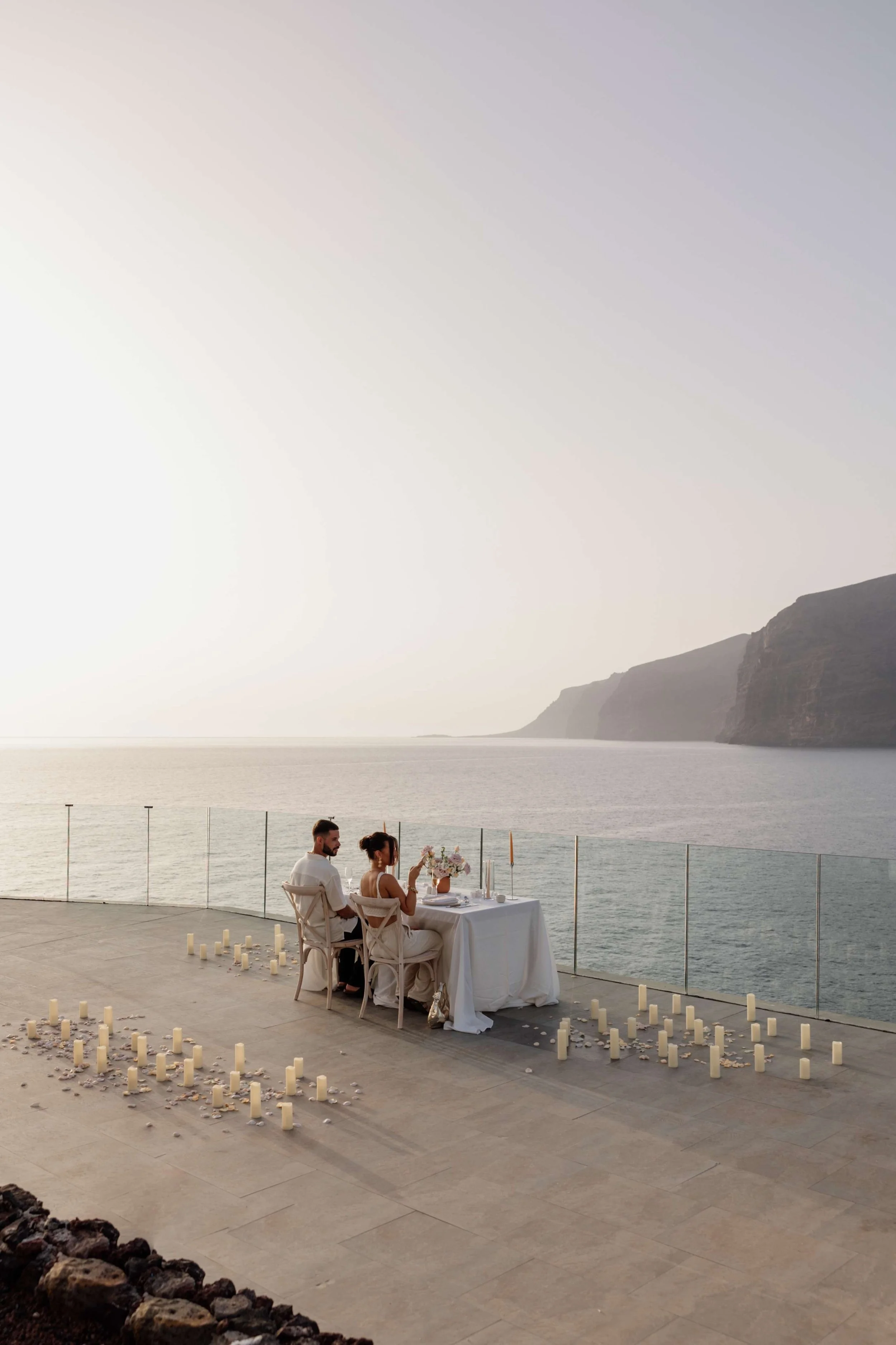 A couple having dinner at a table on an outdoor terrace by the sea, with candles and flower decorations, during sunset.