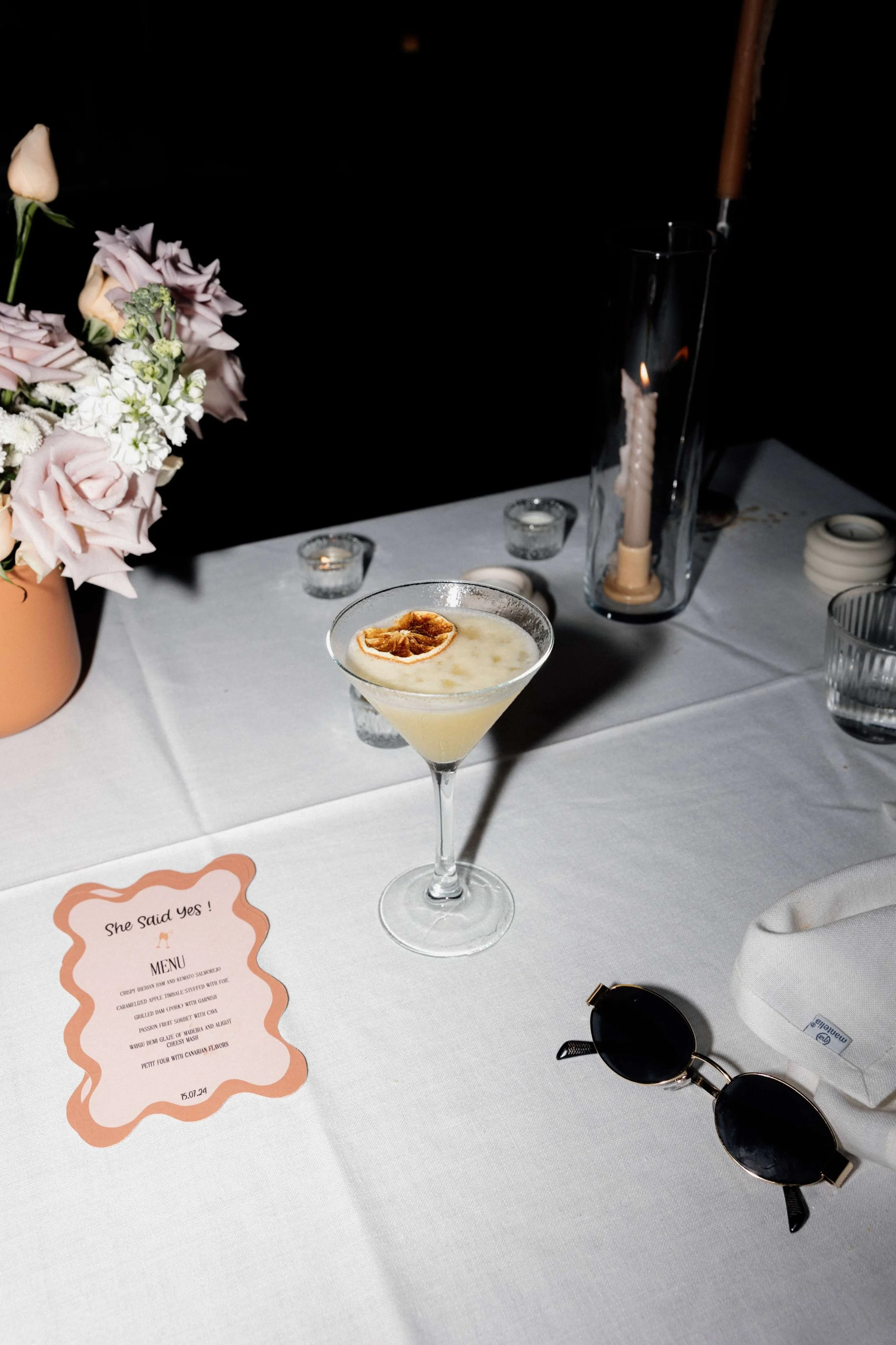 A table set for a celebration with a cocktail drink topped with a dried fruit slice, a pink flower arrangement, candles, a menu, sunglasses, and a cloth napkin.
