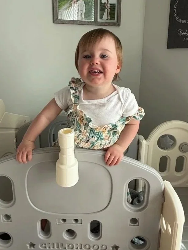 A young girl standing behind a baby hospital bed with a rolled-up paper towel on top of it, smiling at the camera in a room with framed photos on the wall.